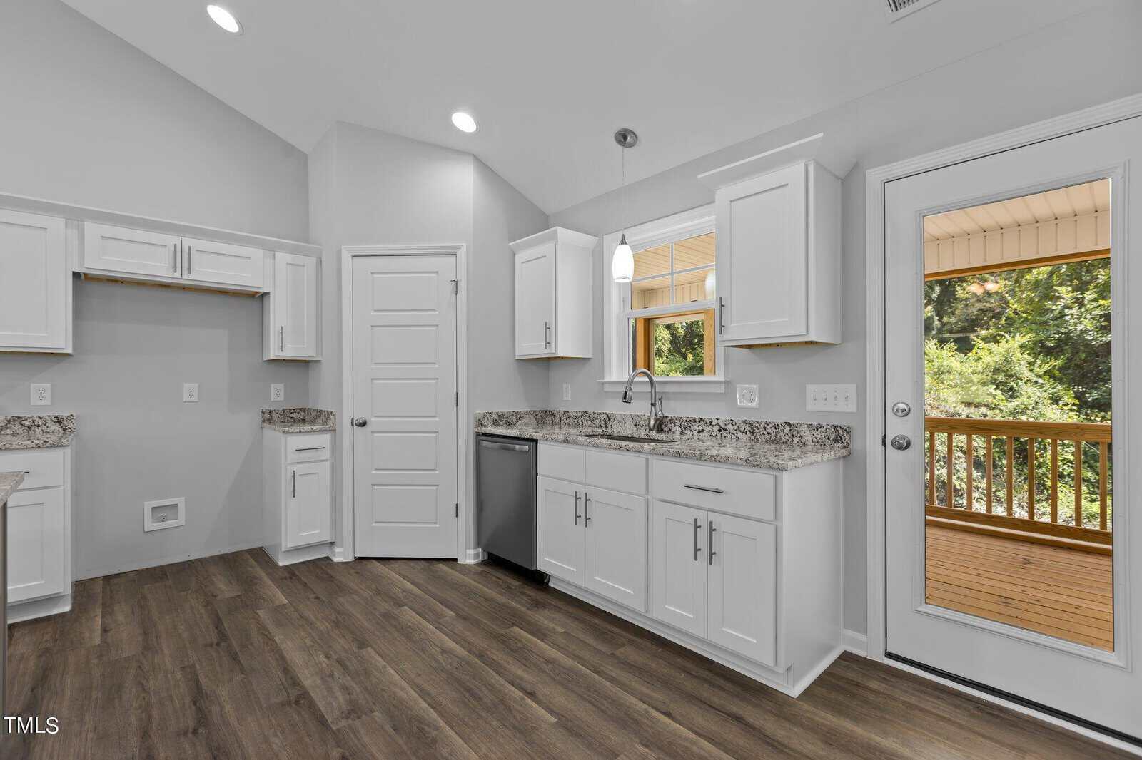Bright kitchen with white cabinets, granite countertops, stainless steel dishwasher, wood flooring, a window over the sink, and a glass door leading to a covered wooden deck with greenery outside.