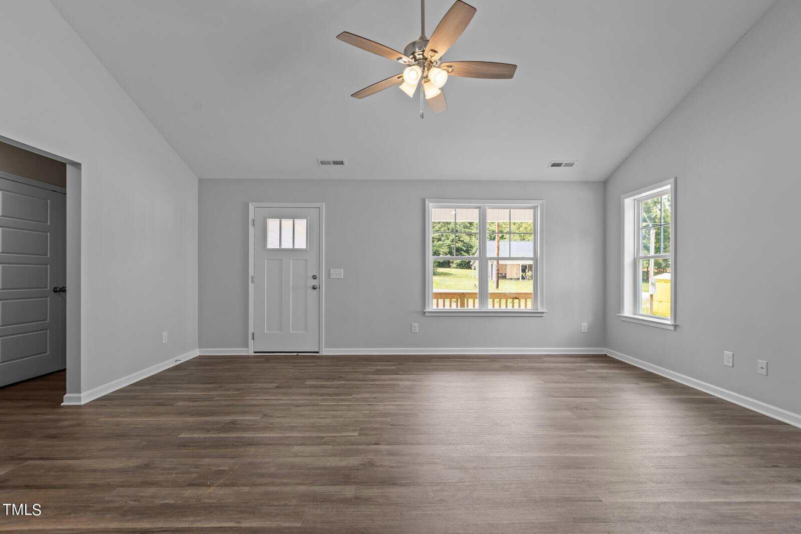Empty, modern living room with gray walls, wood flooring, ceiling fan with lights, white trim, door, and two large windows letting in natural light, offering a view of a porch and greenery outside.