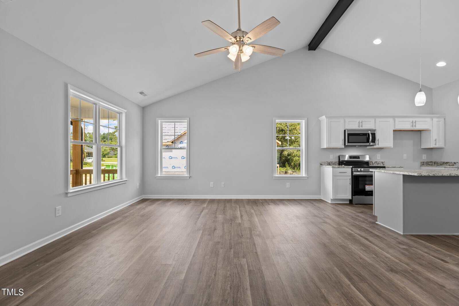 Bright, empty open-concept living space with wood floors, vaulted ceiling, ceiling fan, large windows, and a modern kitchen with white cabinets and granite countertops. Natural light fills the room.