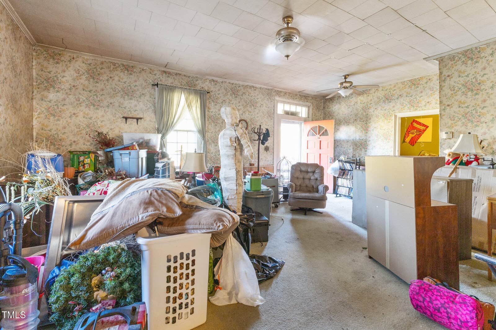 A cluttered living room with scattered boxes, laundry baskets, various furniture, a mannequin, and decor items. Light filters through windows with sheer curtains, and the front door stands open.