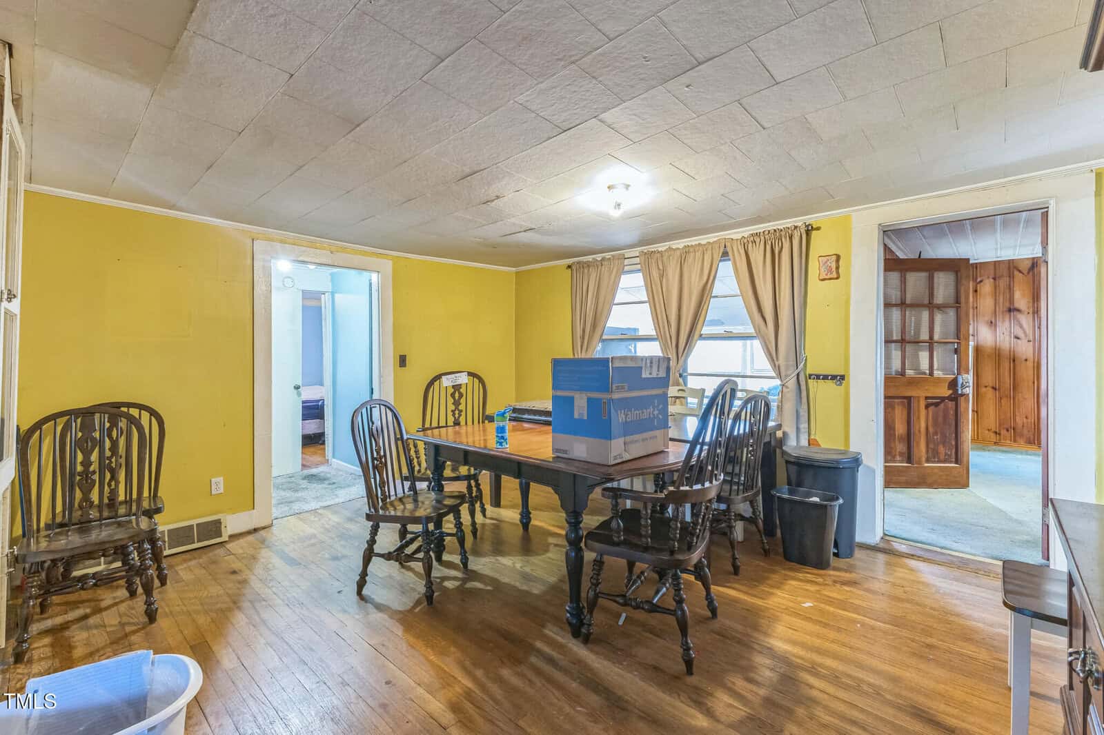 A dining room with yellow walls, a wooden table, six chairs, and boxes on the table. The room has hardwood floors, light-colored ceiling tiles, and multiple doorways leading to adjacent rooms. Brown curtains cover the windows.