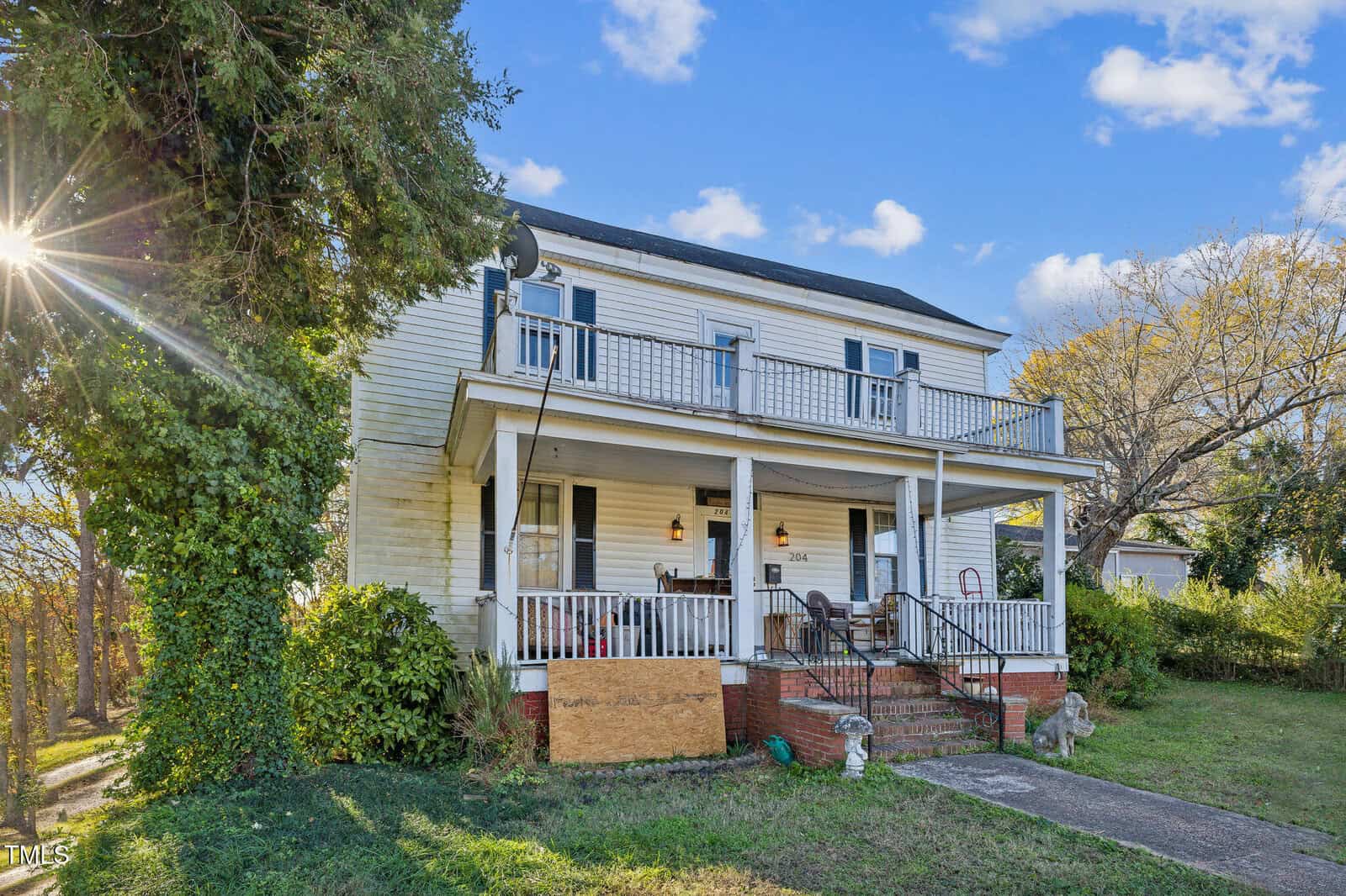 A two-story white house with a covered front porch, black shutters, and a balcony. The porch has chairs and a boarded-up section. There is greenery around the house and sunlight peeking through the trees.