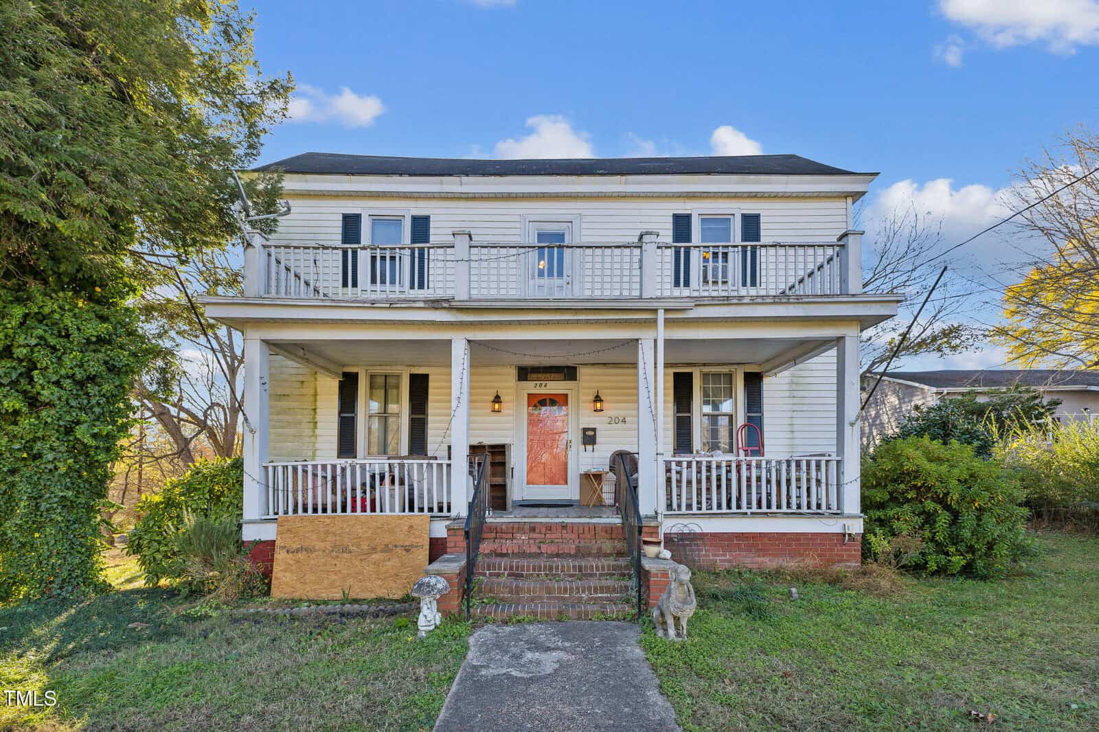 A two-story white wooden house with two upper balconies, a central front door, two porch columns, brick steps, and overgrown grass. There is a boarded-up section on the lower left porch and two small statues by the steps.