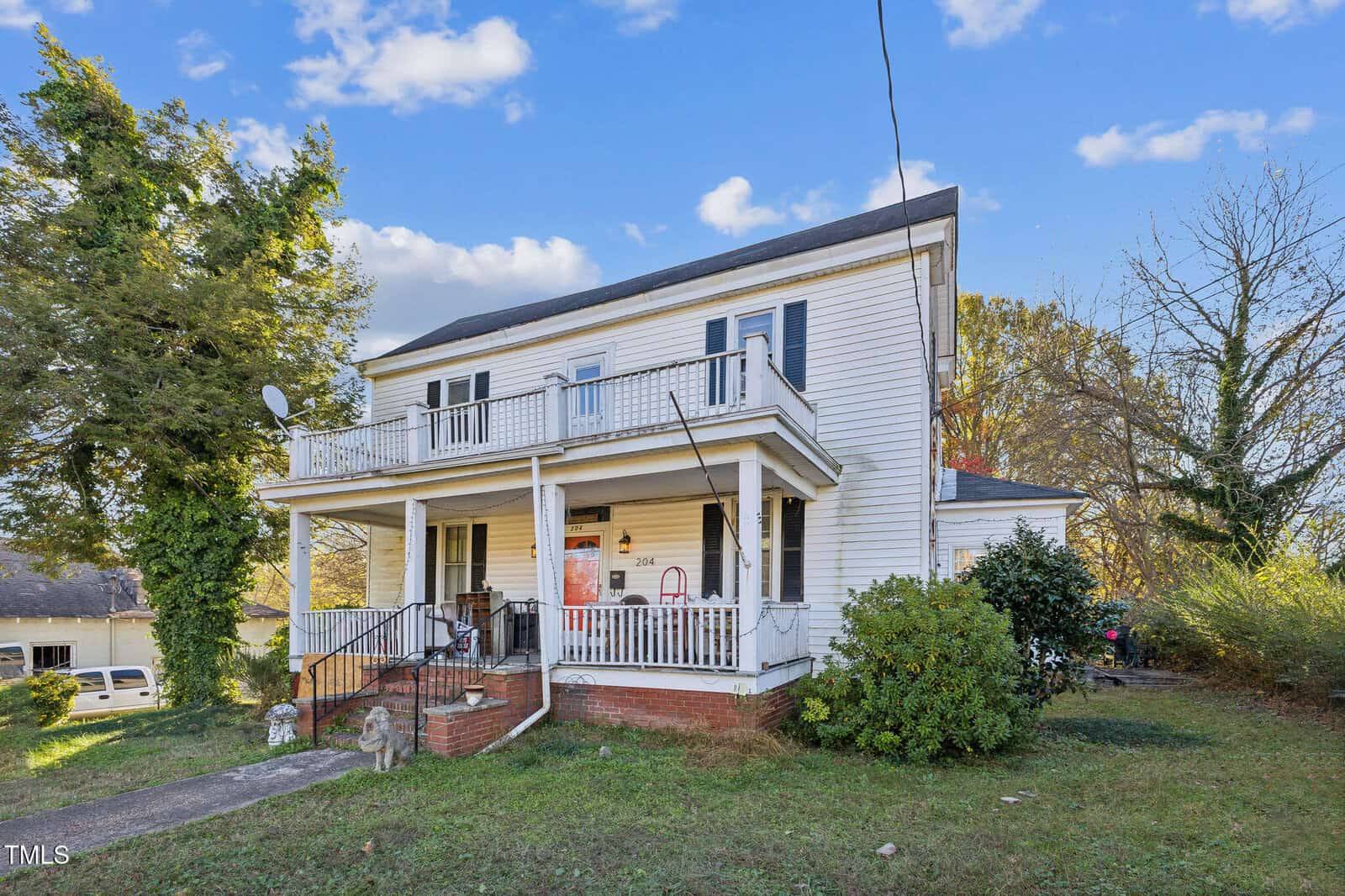 Two-story white house with a covered front porch, black shutters, and an upper balcony. The yard has grass, shrubs, and some overgrown greenery. A concrete walkway leads to steps up to the porch.