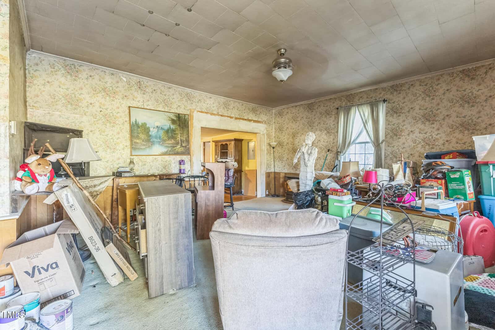 A cluttered living room with mismatched furniture, boxes, and various items scattered around. The walls have floral wallpaper, and a ceiling fan hangs above. Sunlight enters through a window with sheer curtains.