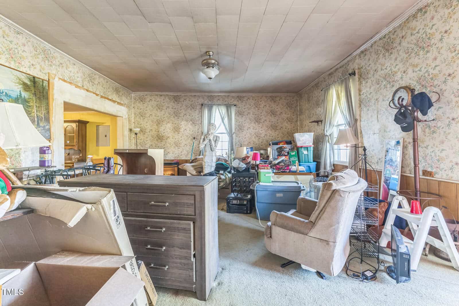 A cluttered living room filled with boxes, bins, and furniture. Floral wallpaper, a ceiling fan, and natural light from several windows are visible. The space appears in need of organization and cleaning.