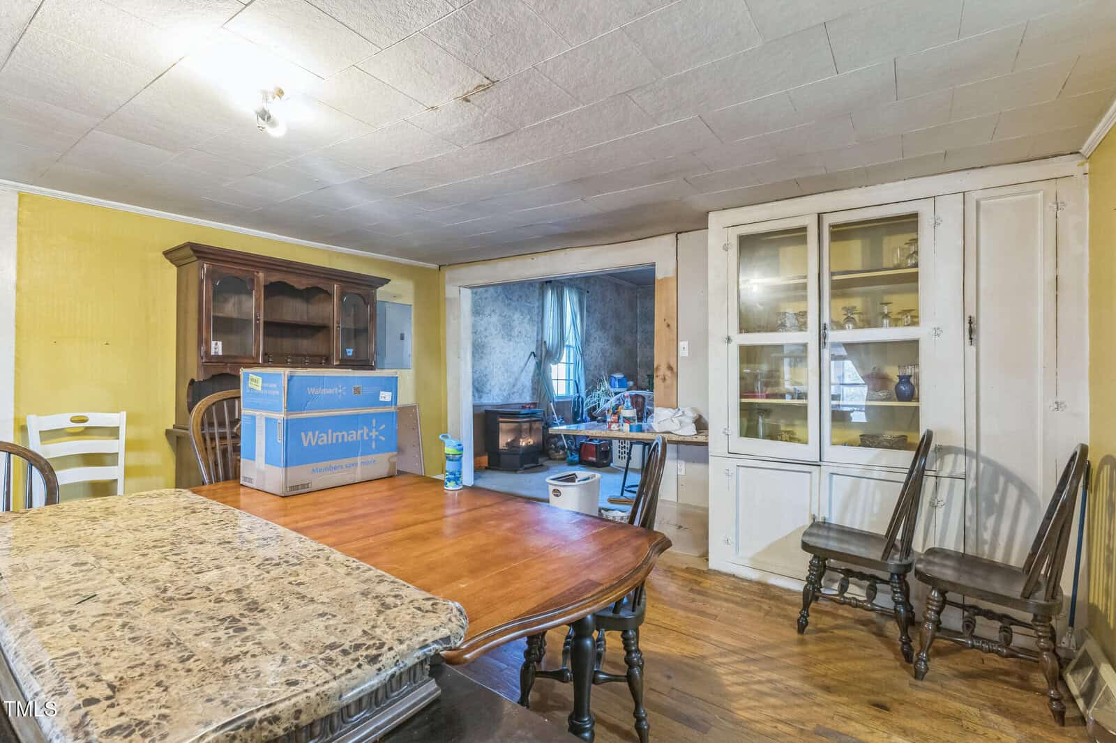 A dining room with a wooden table, several chairs, stacked boxes, and a cabinet with glass doors. An open doorway shows a living room with a wood stove, chairs, and various household items. The ceiling has visible stains.
