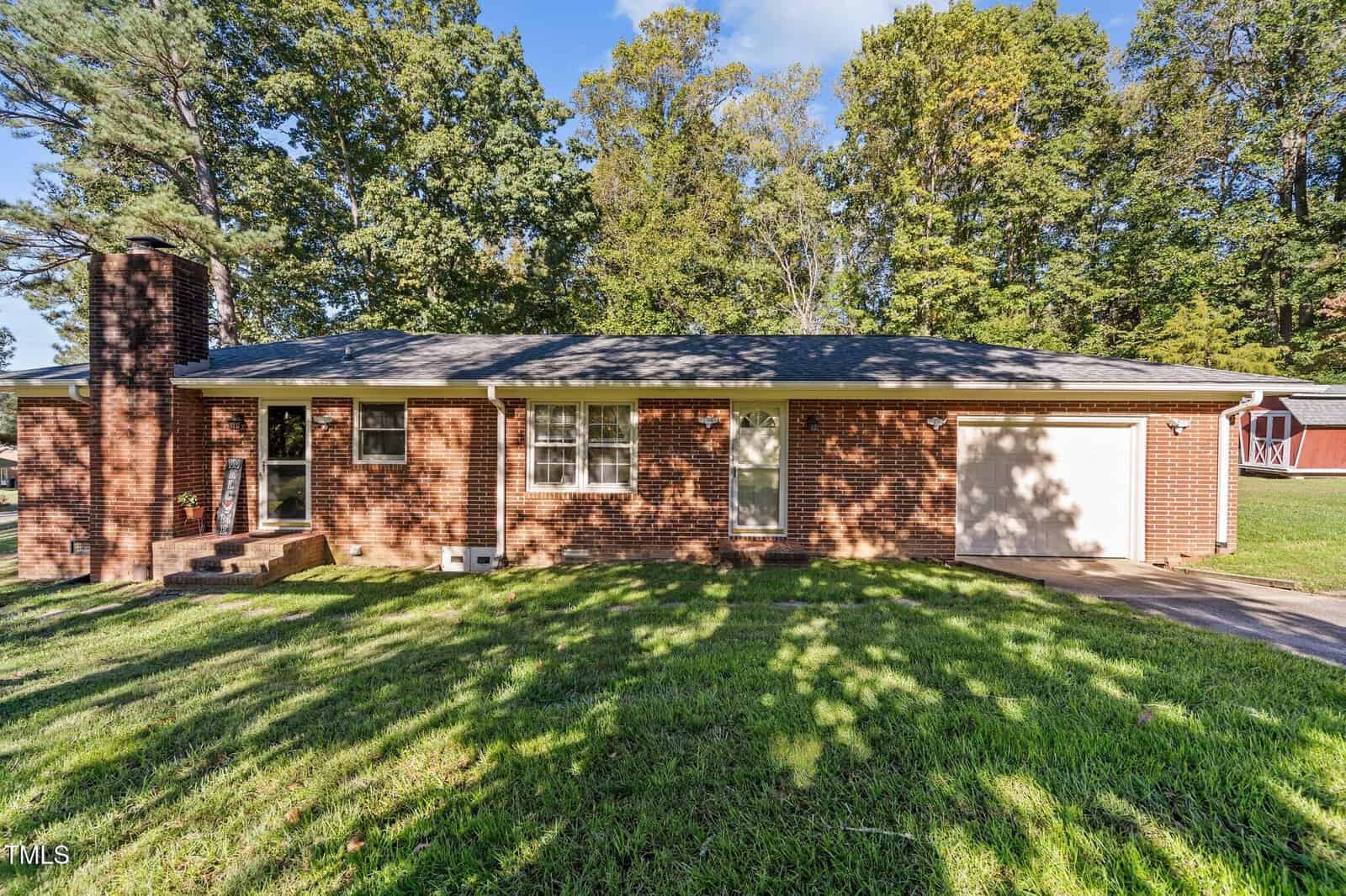 A single-story brick house with a front porch, a chimney, and an attached white garage door. The lawn is grassy with some trees in the background. The sky is clear and sunny.