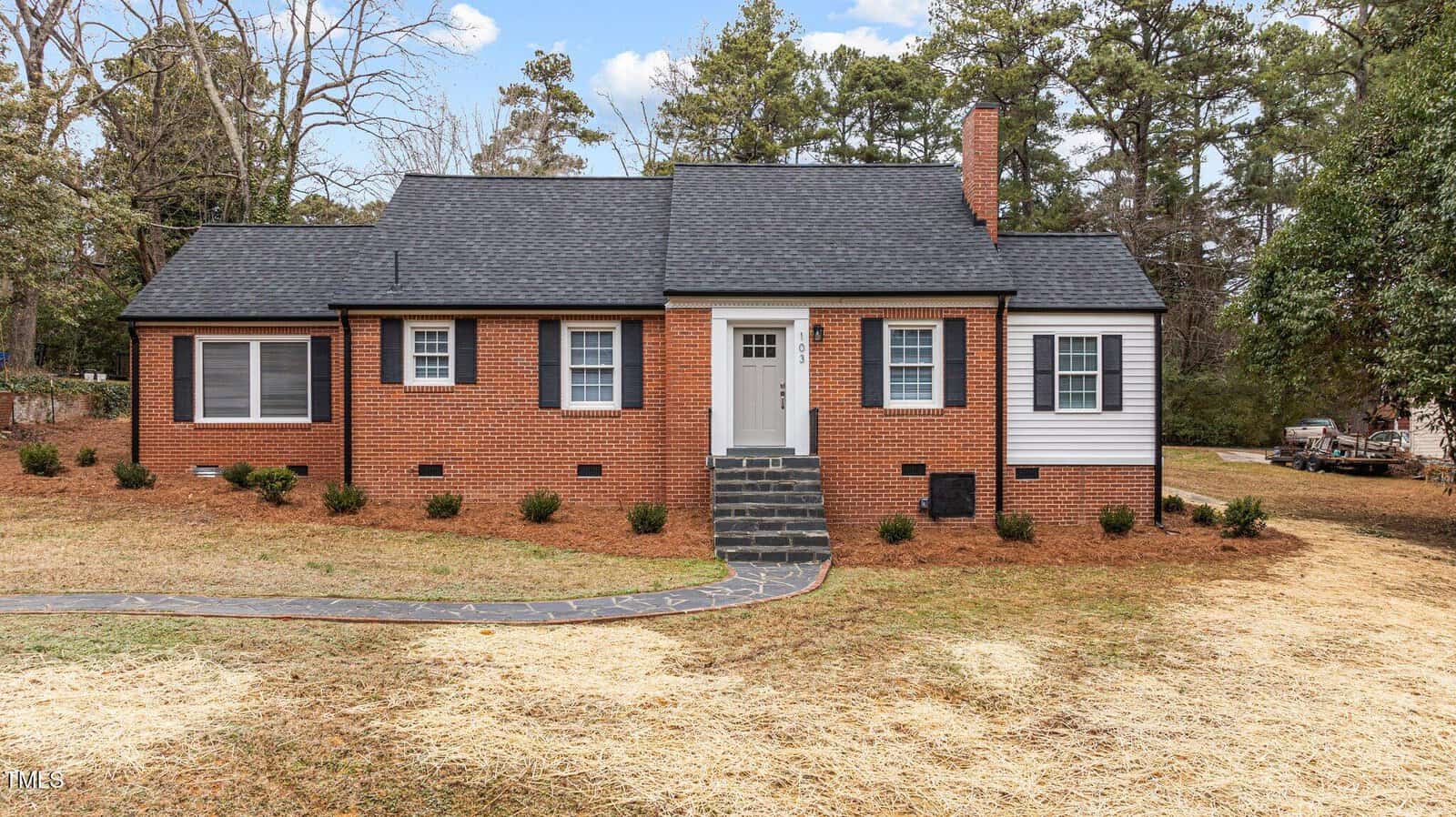 Located at 103 Park Drive, this charming Oxford residence is a single-story brick house with a black shingled roof and white window shutters. A small front porch, complete with steps to the entrance, adds to its appeal, while the yard boasts sparse grass and trees in the background.