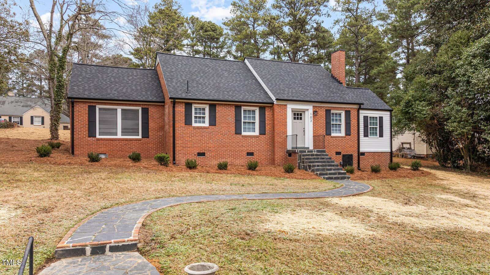 A red brick house with a dark gray roof, white trim, and a chimney stands surrounded by trees. A curved stone path leads to the front door. The yard has some grass and bushes, with other homes visible in the background.