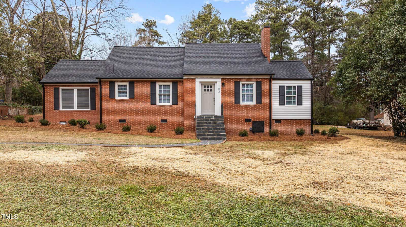 A single-story brick house with a dark roof and a central chimney. The front door is white with a small window, accessed by a short flight of steps. The yard is grassy with newly planted shrubs along the front. Trees are visible in the background.