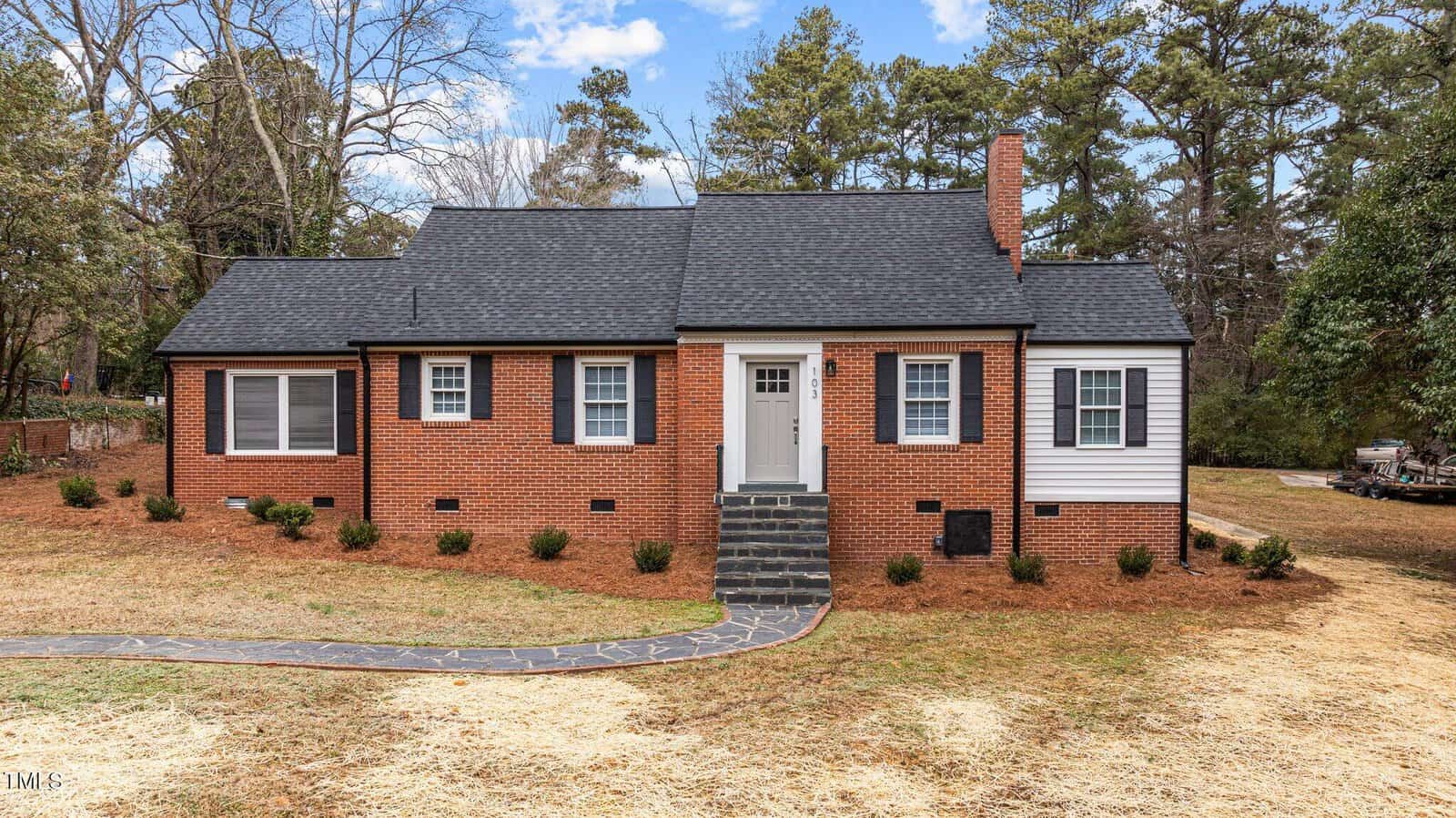 A single-story brick house with black roof shingles, a white front door, and dark window shutters. The house is surrounded by a grassy yard with a paved walkway leading to the entrance. Trees are visible in the background under a partly cloudy sky.