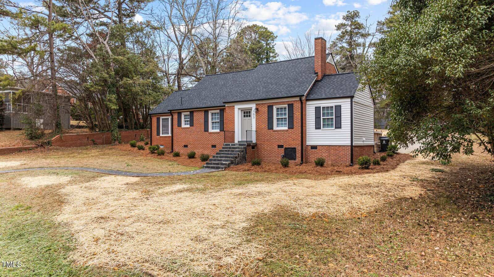 A single-story brick house with white siding and a dark gray roof. There is a small front porch with steps leading up to the entrance. The yard is partially covered in straw, and there are trees and bushes surrounding the house.