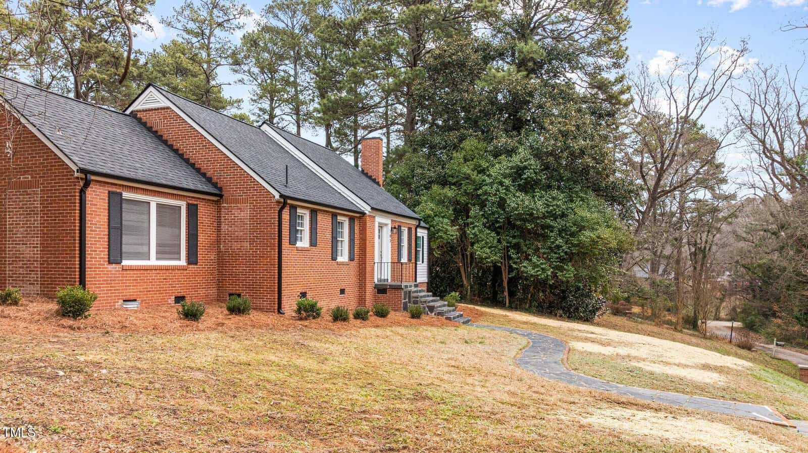 A brick house with a sloped roof, surrounded by grass and trees, is pictured. There are steps leading to the entrance, and the sky is partly cloudy. A path leads from the sidewalk to the front door.