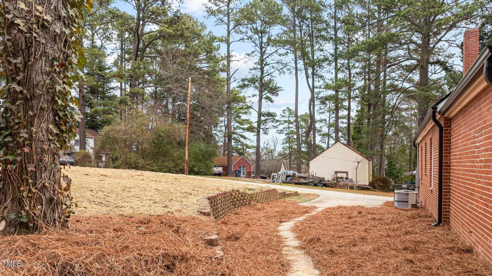 A residential area with a brick house on the right and a mulched pathway leading down. In the background are tall pine trees, a driveway, and additional residential buildings. The sky is partly cloudy.