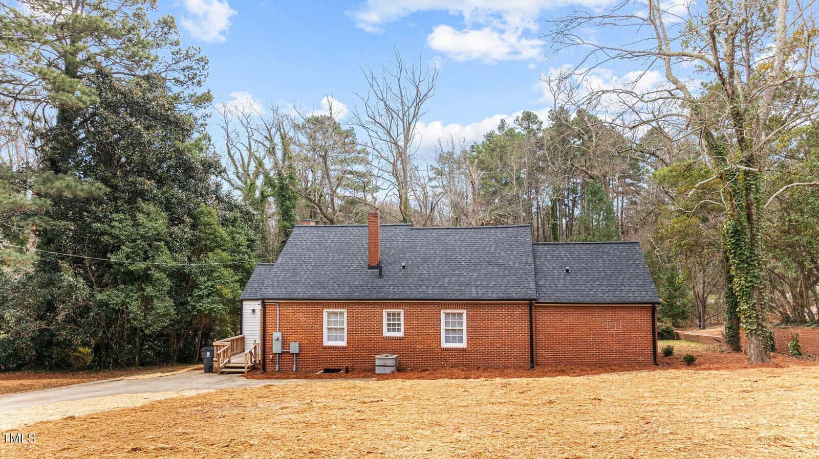 A small, red-brick house with a black shingled roof is surrounded by tall trees. The yard is cleared, with straw covering the ground. Steps lead up to a side door, and utility units are visible at the back of the house. The sky is partly cloudy.