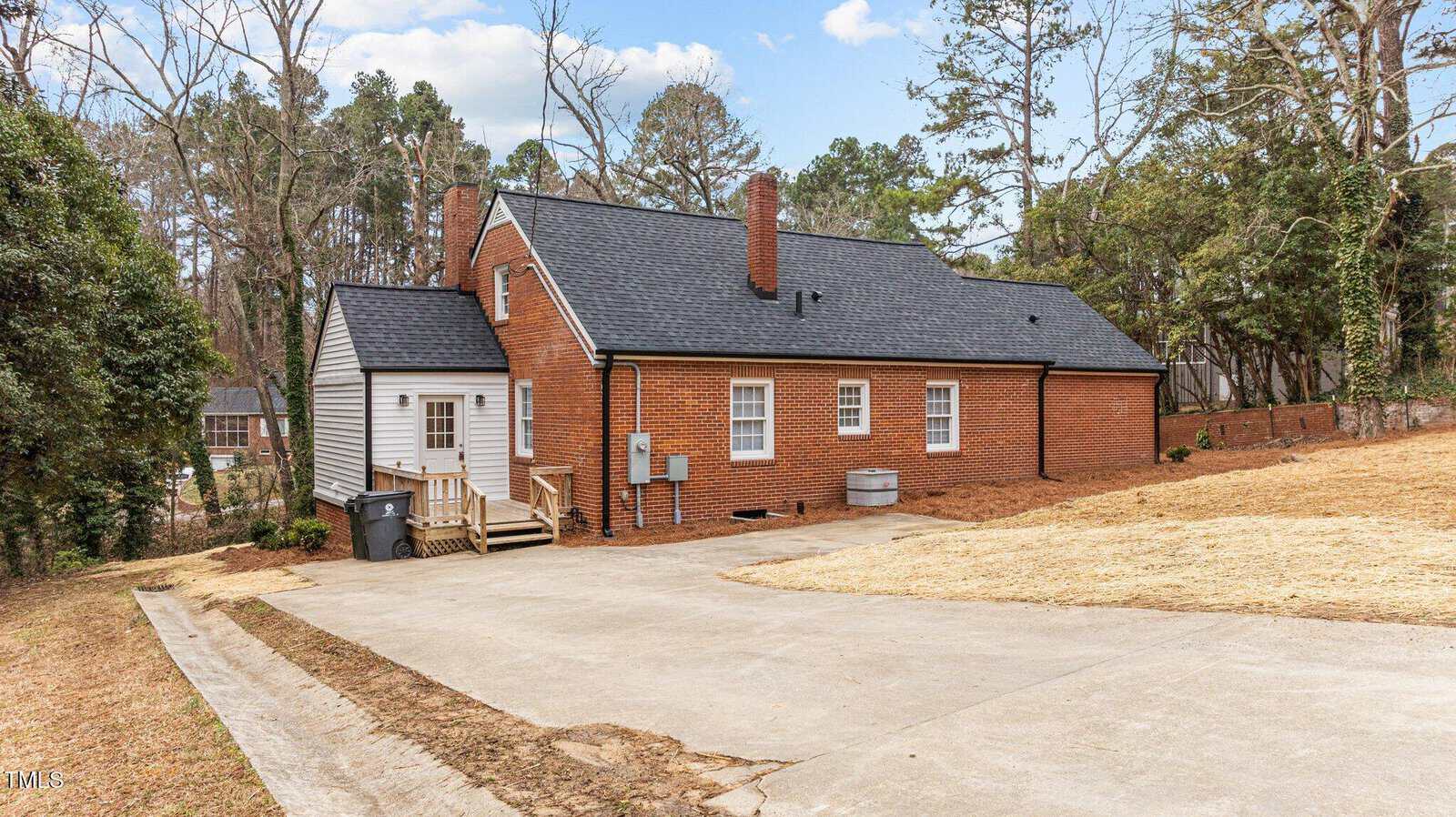 A red brick one-story house with a black shingled roof, featuring a small back porch and a chimney. The yard is mostly bare with dried grass. A driveway leads up to the house, surrounded by trees and shrubs.