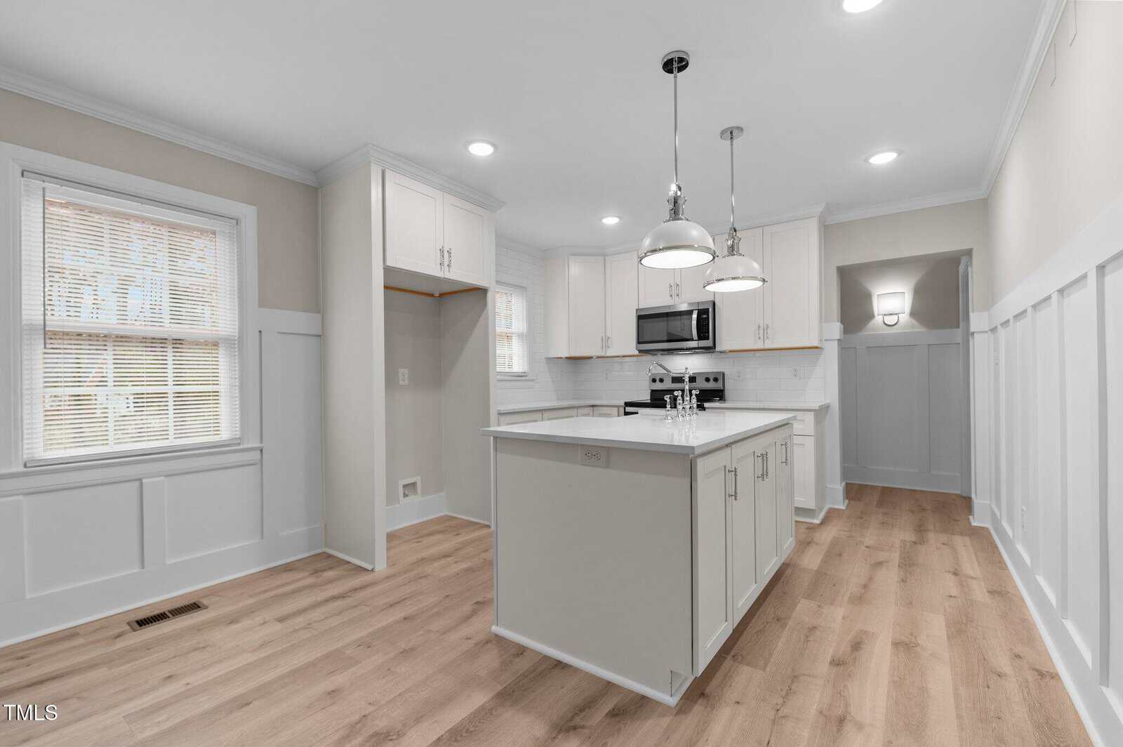 A modern kitchen features white cabinetry, a central island with a light gray countertop, and stainless steel appliances. Pendant lights hang above the island. The floor is light wood, and the walls have white wainscoting and windows with blinds.