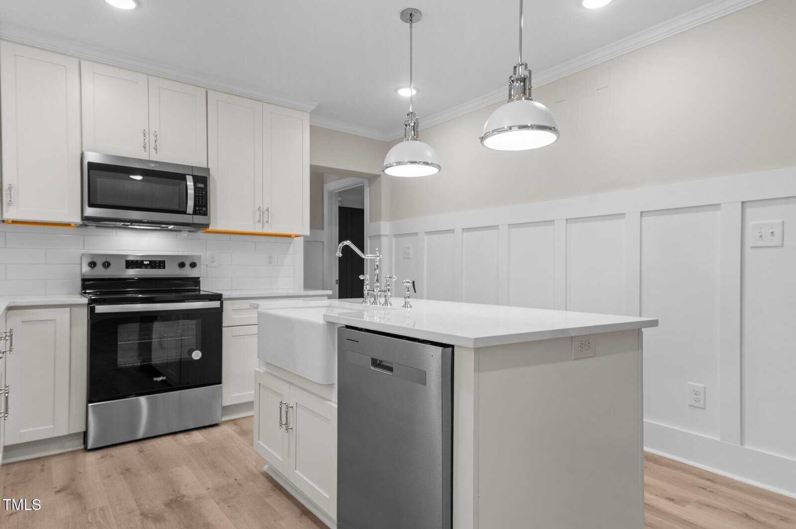 Modern kitchen with light-colored cabinetry and an island featuring a dishwasher and sink. Stainless steel microwave and oven are built into the cabinets. Two pendant lights hang above the island. White backsplash and light wood floors complete the look.
