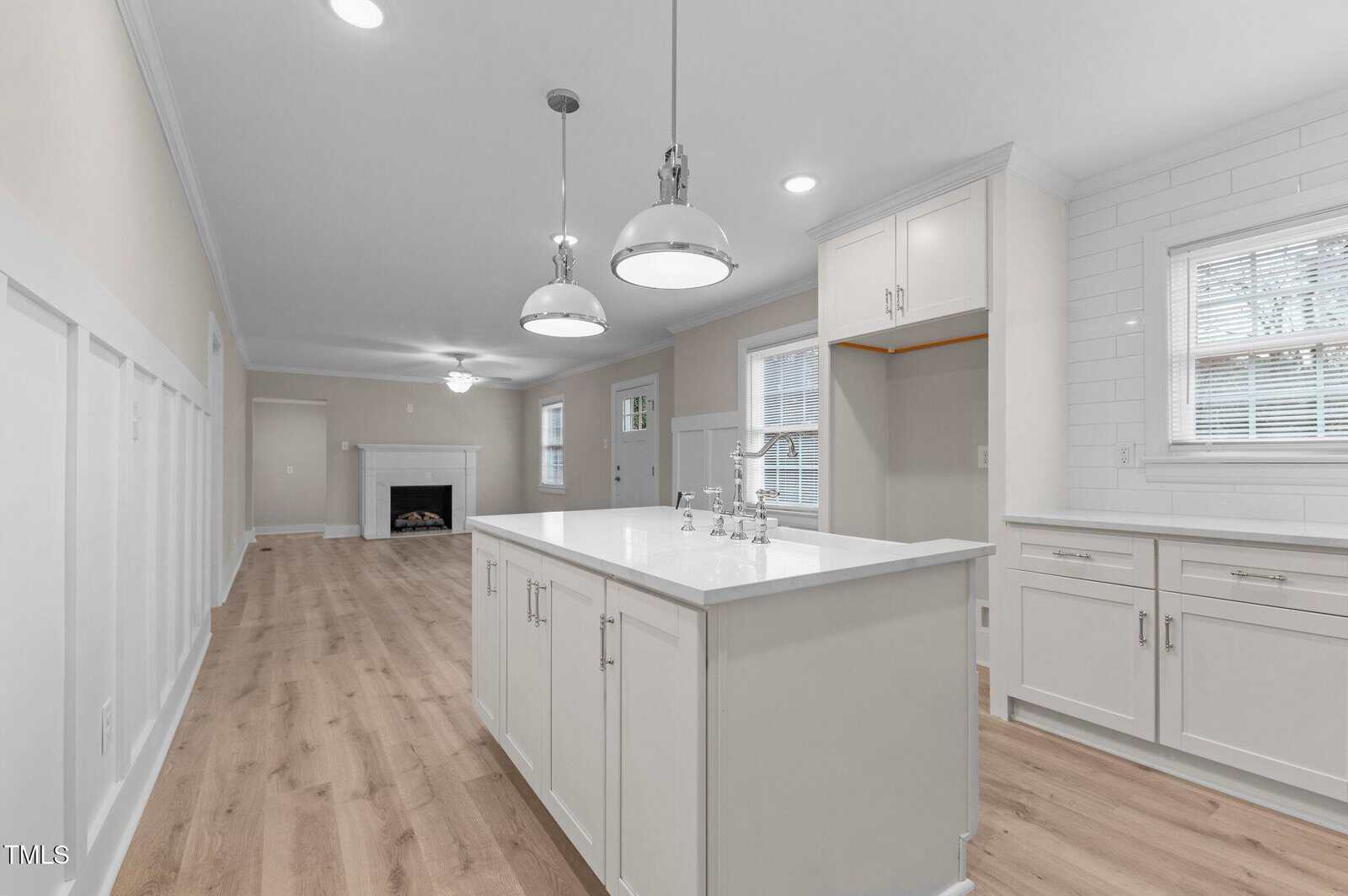 A modern kitchen with white cabinets and an island featuring a sink and chrome faucet. Two pendant lights hang above the island. The kitchen opens into a living area with a fireplace. Light wood flooring runs throughout the space.