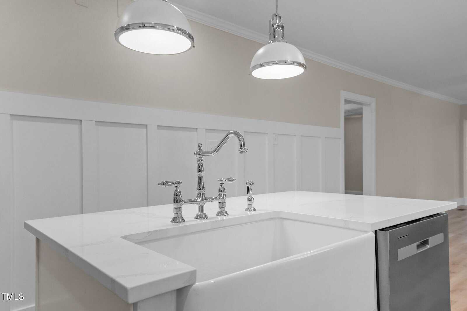 A modern kitchen sink with a high-arch faucet is set into a white countertop. Two pendant lights hang above, illuminating the space. In the background, a light-colored wall features subtle decorative molding. A dishwasher is installed to the right.