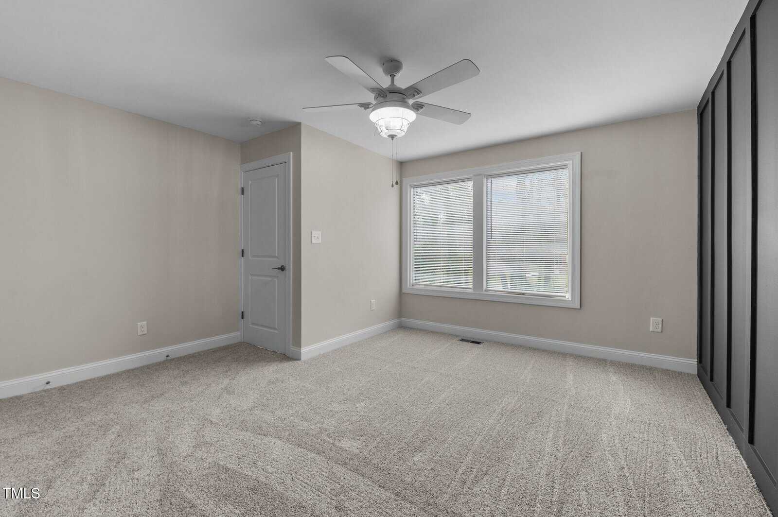 An empty room with light beige walls and carpeted floor. A ceiling fan with a light hangs from the middle of the ceiling. A window with blinds is on the right wall, and a closed door is on the left.