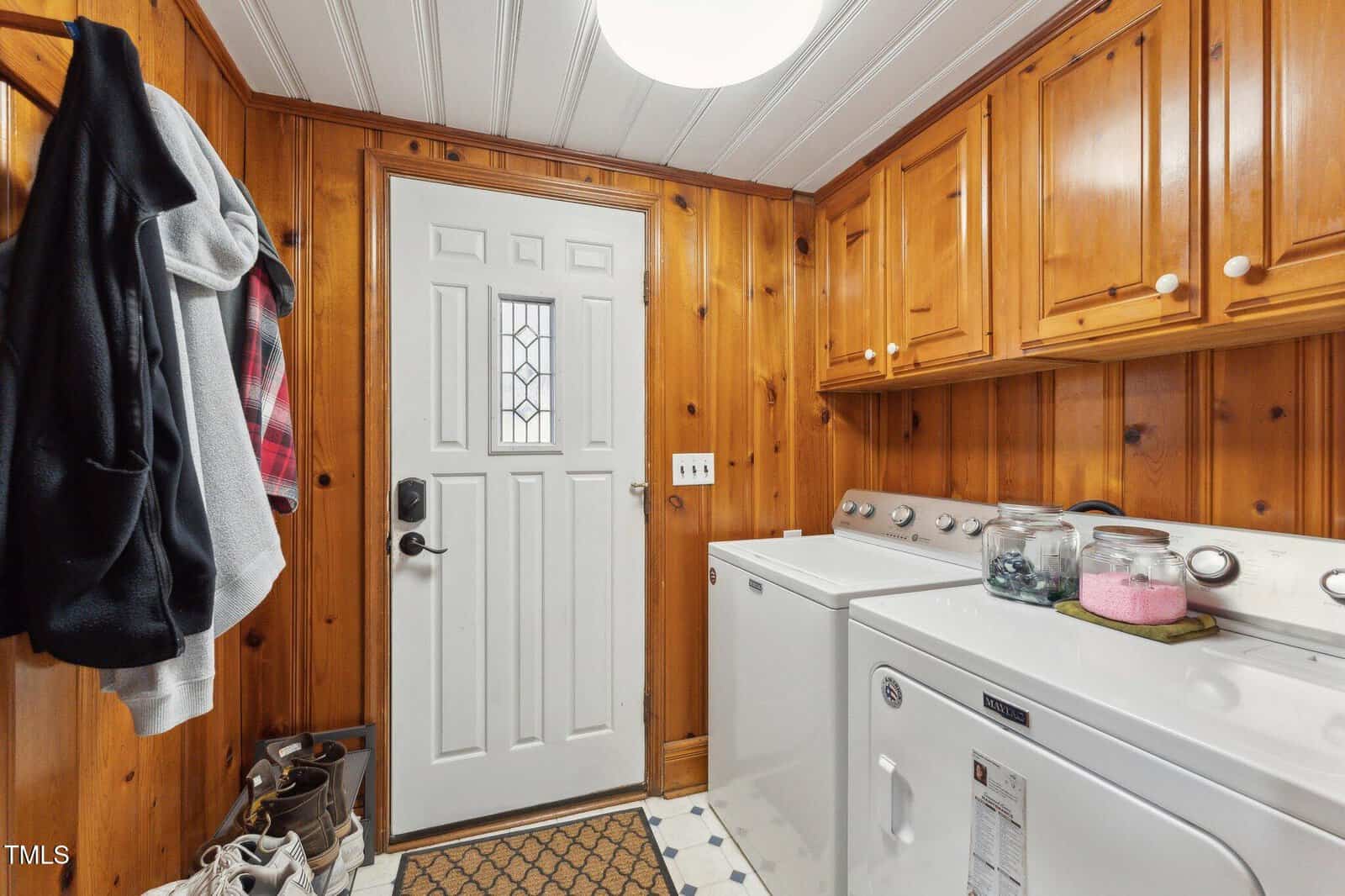 A laundry room with wooden paneled walls, a white door with a small window, a washer and dryer, overhead cabinets, a coat rack with jackets, and shoes on a small mat.