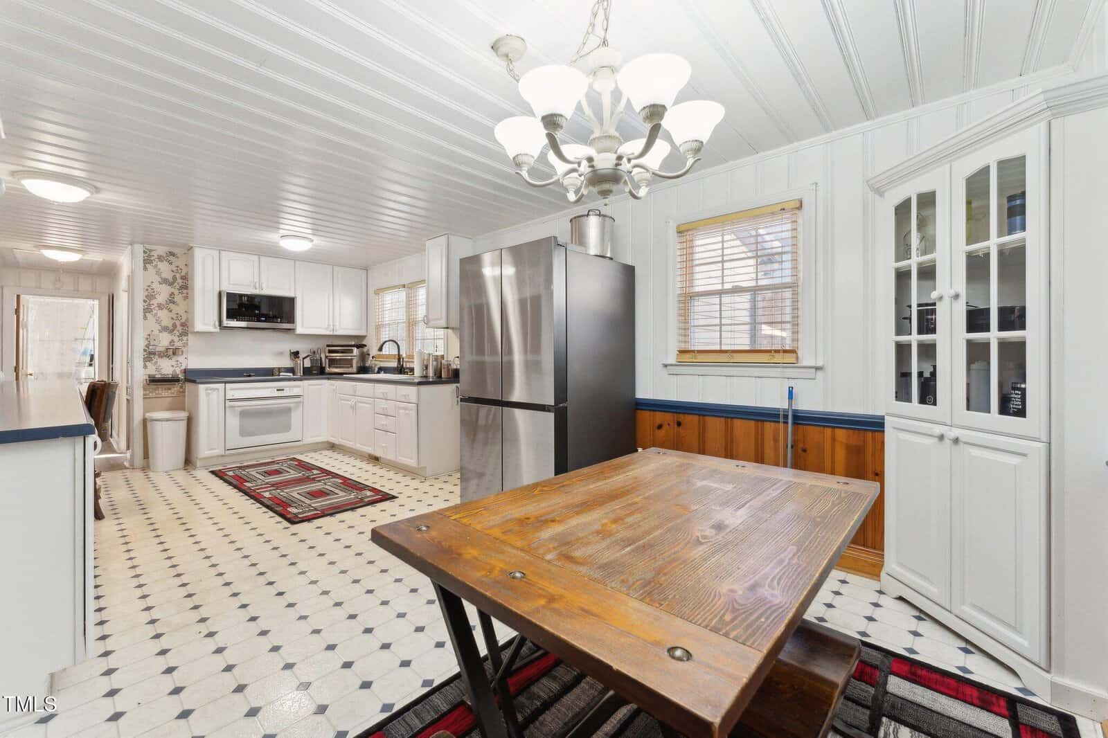 A spacious kitchen with white cabinets, stainless steel appliances, a wooden dining table, glass-front cabinet, patterned rugs, and a chandelier, featuring white paneled walls and ceiling, and two windows with blinds.