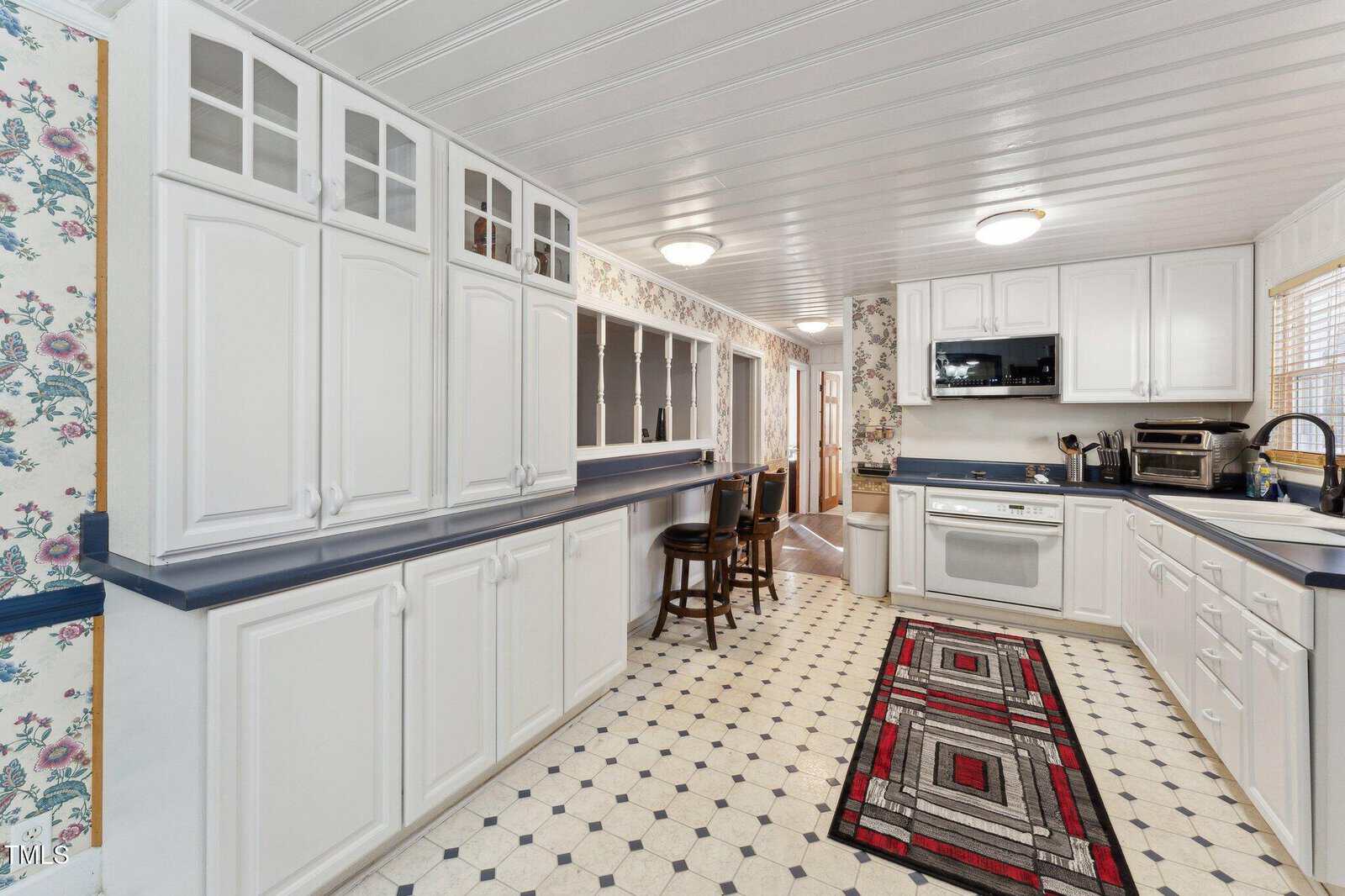 A kitchen with white cabinets, blue countertops, a patterned black, white, and red rug, white appliances, and floral wallpaper. A window is above the sink, and two barstools are at a counter near the far wall.