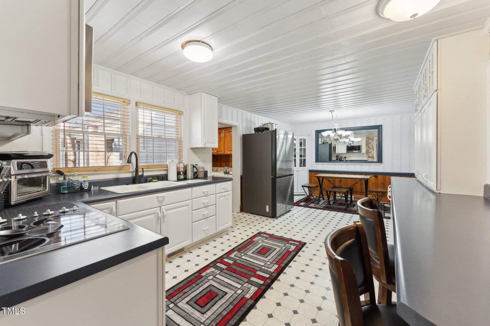Bright kitchen with white cabinets, dark countertops, a gas stove, black refrigerator, and geometric rugs. A dining area with wooden table and chairs is visible in the background under a modern chandelier.