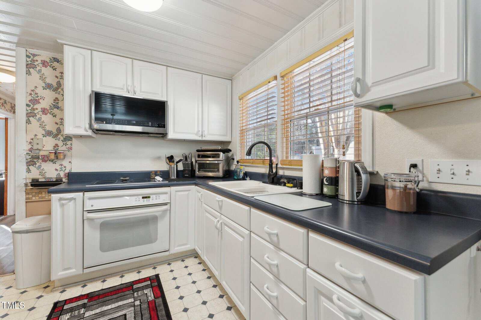 Bright kitchen with white cabinets, black countertops, and white appliances. Large windows with blinds above the sink, and a black-and-red rug on patterned tile flooring. Various small appliances and utensils are on the counters.