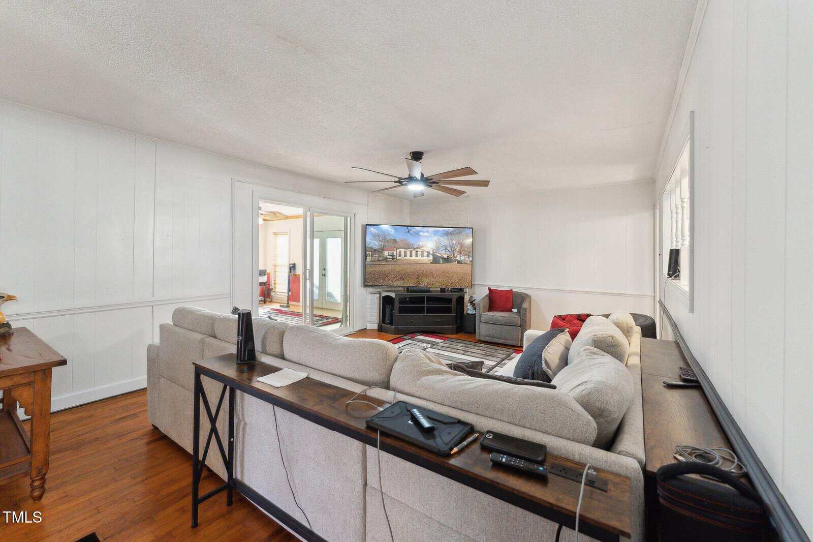 Living room with a large gray sectional, wall-mounted TV, wooden floors, ceiling fan, and neutral walls. The space features a console table behind the sofa and a view into another room through glass doors.