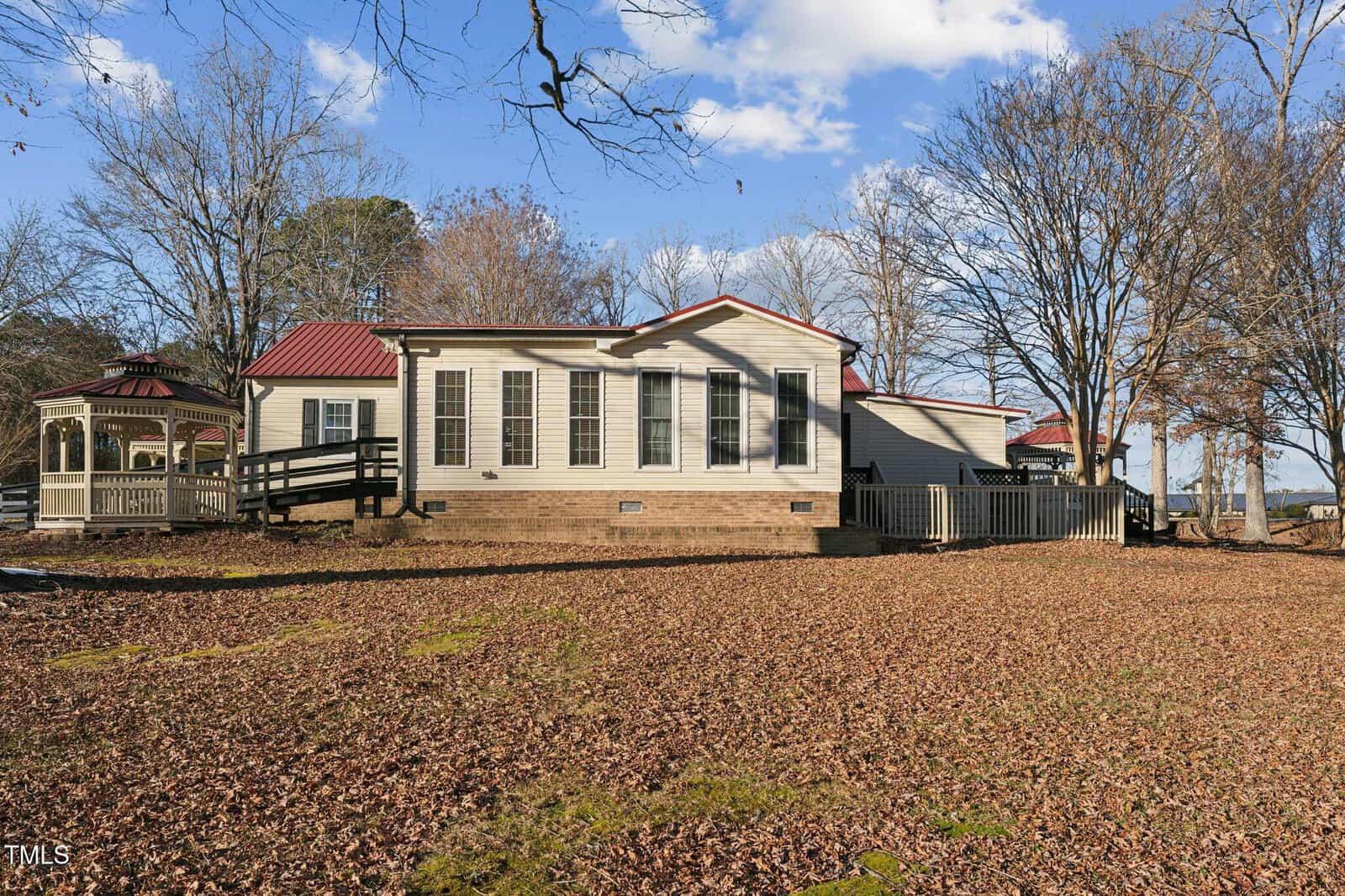 A single-story house with a red metal roof, multiple windows, and a wooden ramp on the left. A gazebo is nearby, and leaf-covered ground with scattered trees surrounds the property under a blue sky.
