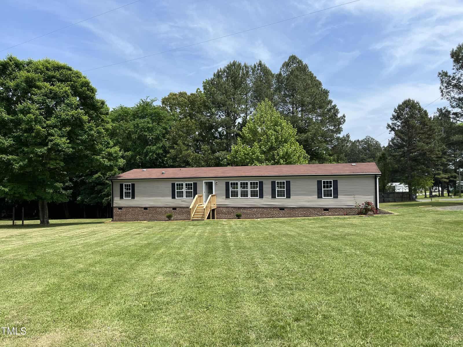 A single-story gray house with a red roof sits on a large, neatly mowed lawn. It has steps leading to the front entrance and several windows. Surrounding trees frame the property under a clear blue sky.