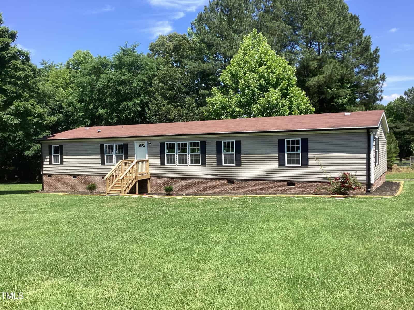 A single-story manufactured home with a brown shingle roof is set on a spacious lawn. The exterior is beige with brick skirting, and there is a small wooden staircase leading to the front door. Tall trees and greenery surround the house in the background.