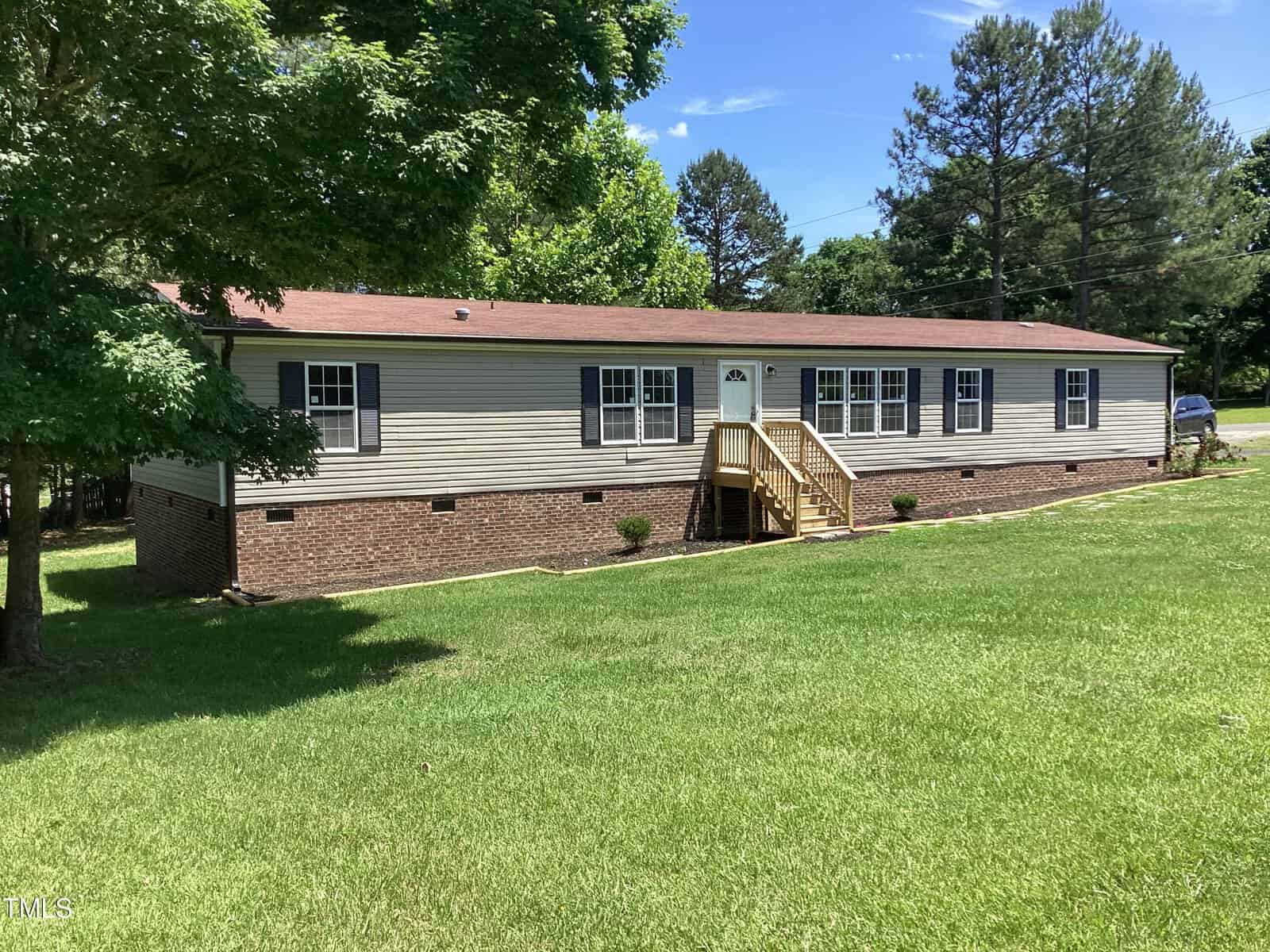 A single-story house with beige siding and a red-brown roof is surrounded by a grassy lawn. It features several windows and a wooden staircase leading to the front door. Tall trees are visible in the background under a clear blue sky.