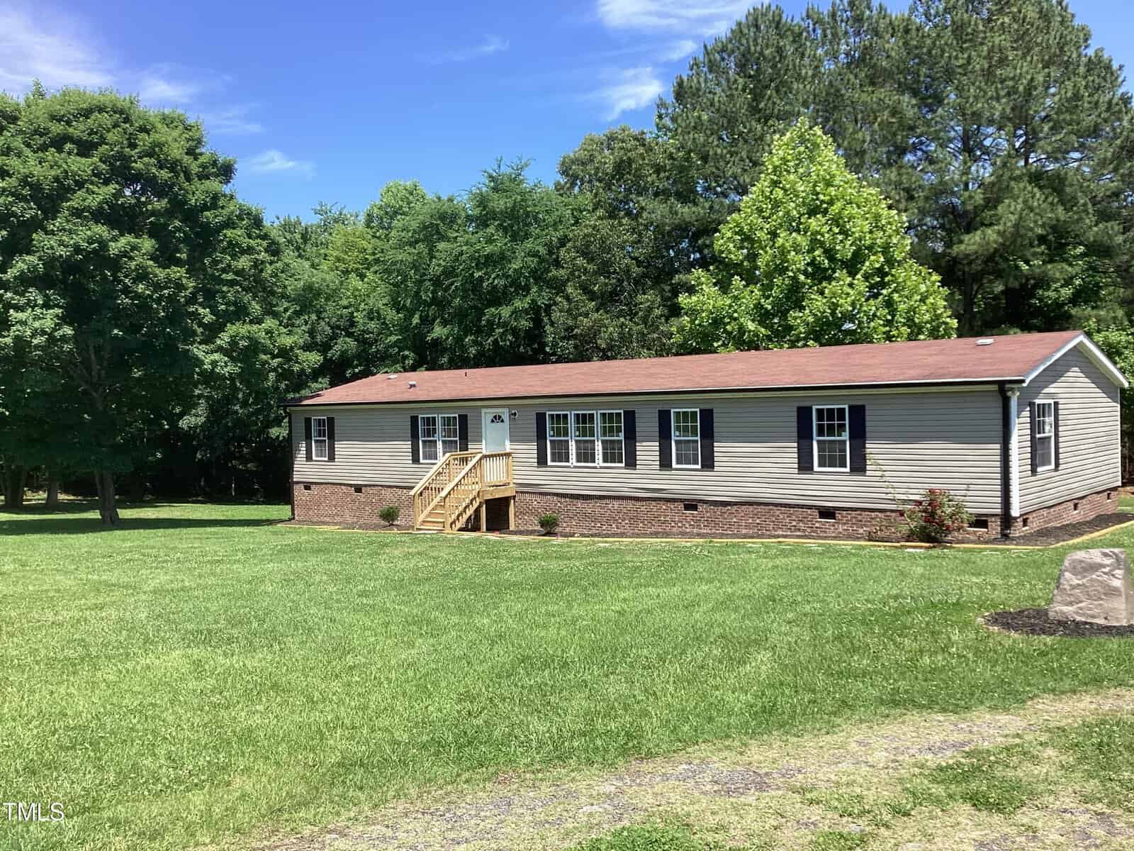 A single-story manufactured home with a brown shingle roof and beige siding is set on a large green lawn. The house has multiple windows and a wooden front porch. In the background, there are trees and a clear blue sky.