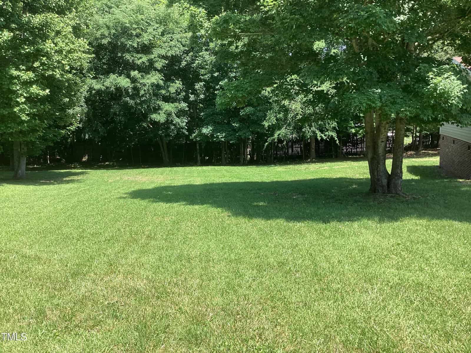 A grassy yard with a well-maintained lawn is bordered by tall, leafy trees. Shadows from the trees extend across the grass. A small portion of a brick structure is visible in the right corner of the image.