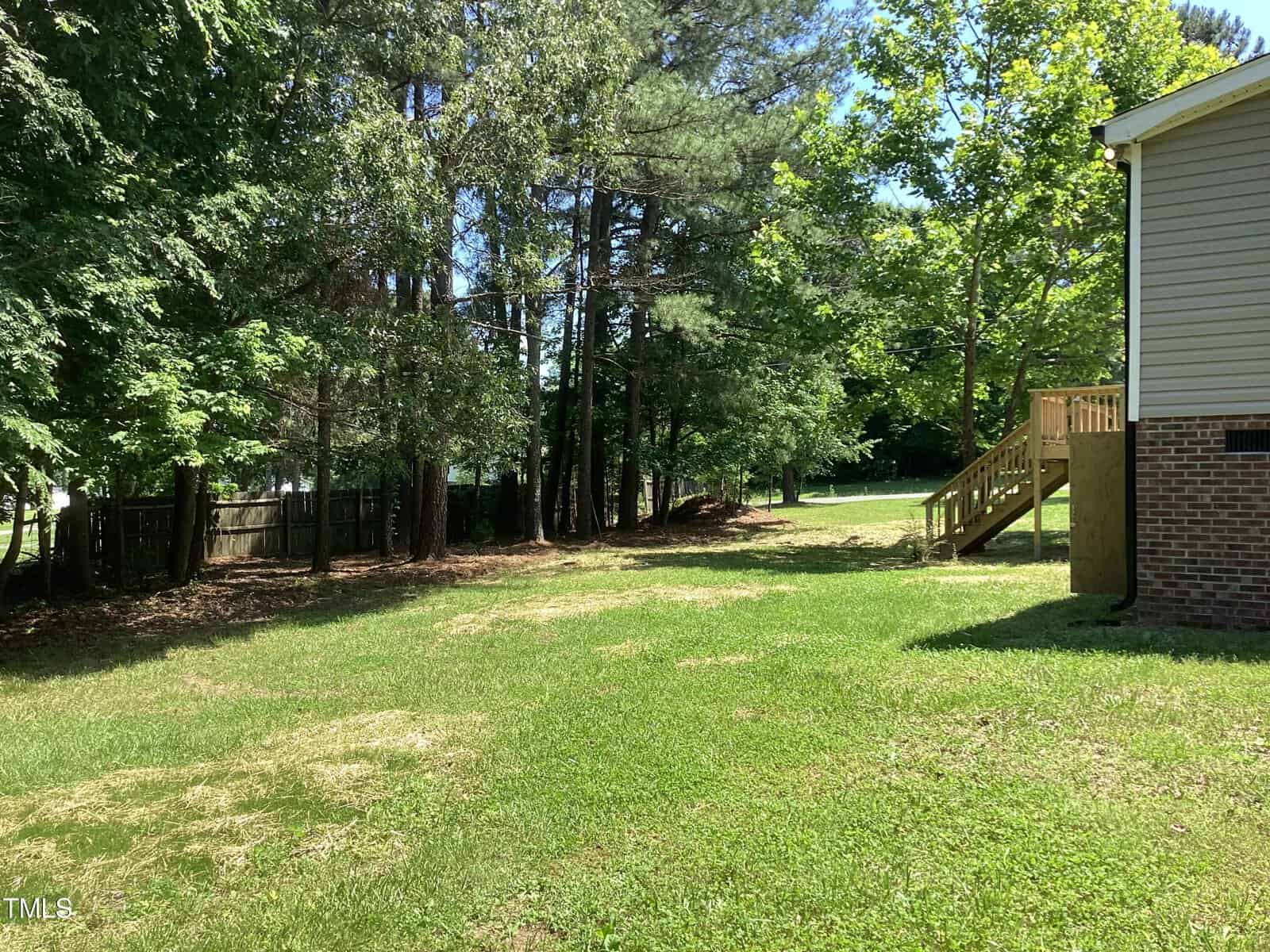 A grassy backyard with a few scattered patches of dirt. Tall trees and a wooden fence line the left side. On the right, there is a brick house with wooden steps leading to the ground. The sky is clear and sunny.