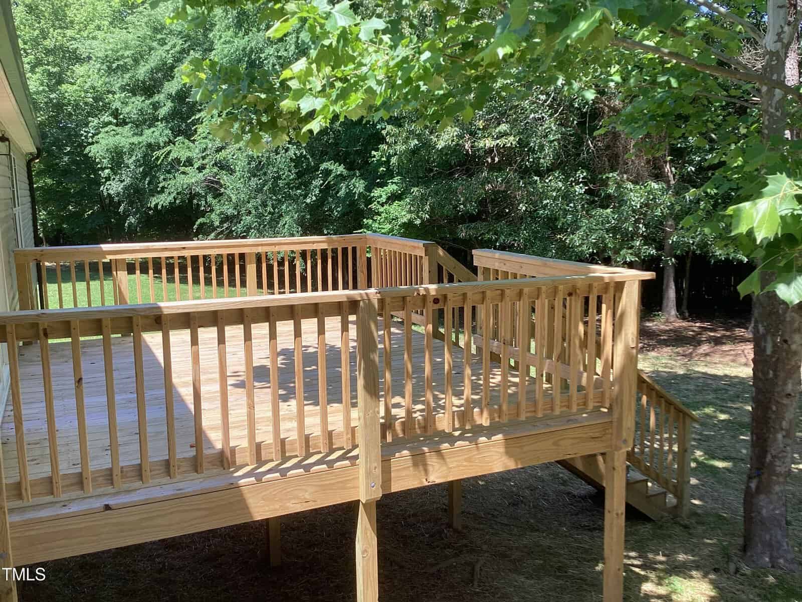 A wooden deck with railings, attached to a house, surrounded by trees and grass. Stairs on the right lead down to the ground. Sunlight filters through the leaves, casting shadows on the deck.