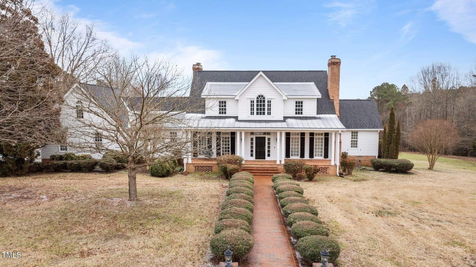A large two-story white house at 1200 Spring Valley Lake Road, Henderson, features a covered front porch, black roof, and two brick chimneys, set on a spacious lawn with trimmed hedges and a brick walkway leading to the entrance.