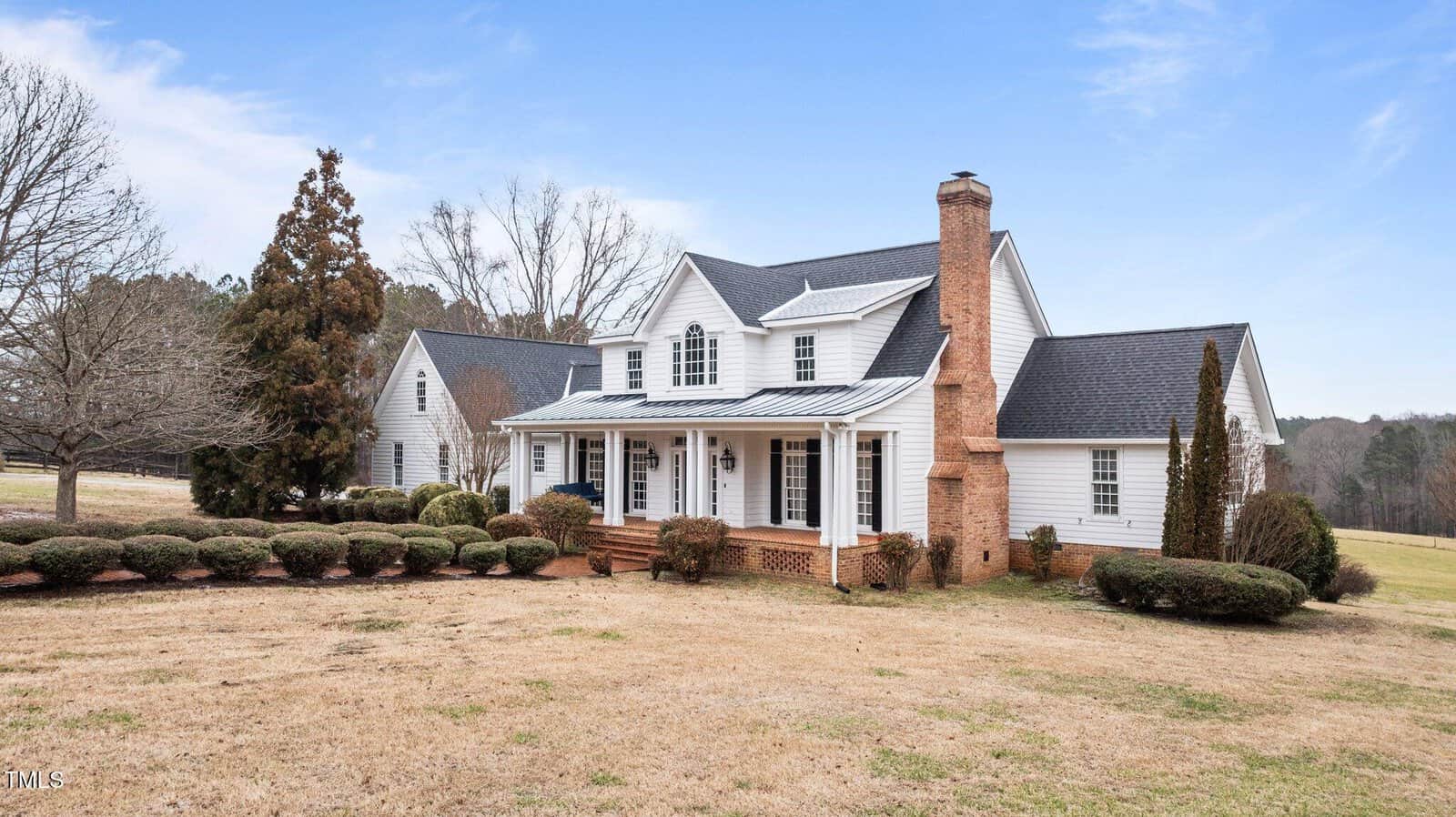 White two-story house with a front porch, brick steps, and a large brick chimney, surrounded by trimmed bushes and trees on a spacious, grassy lawn under a clear blue sky.