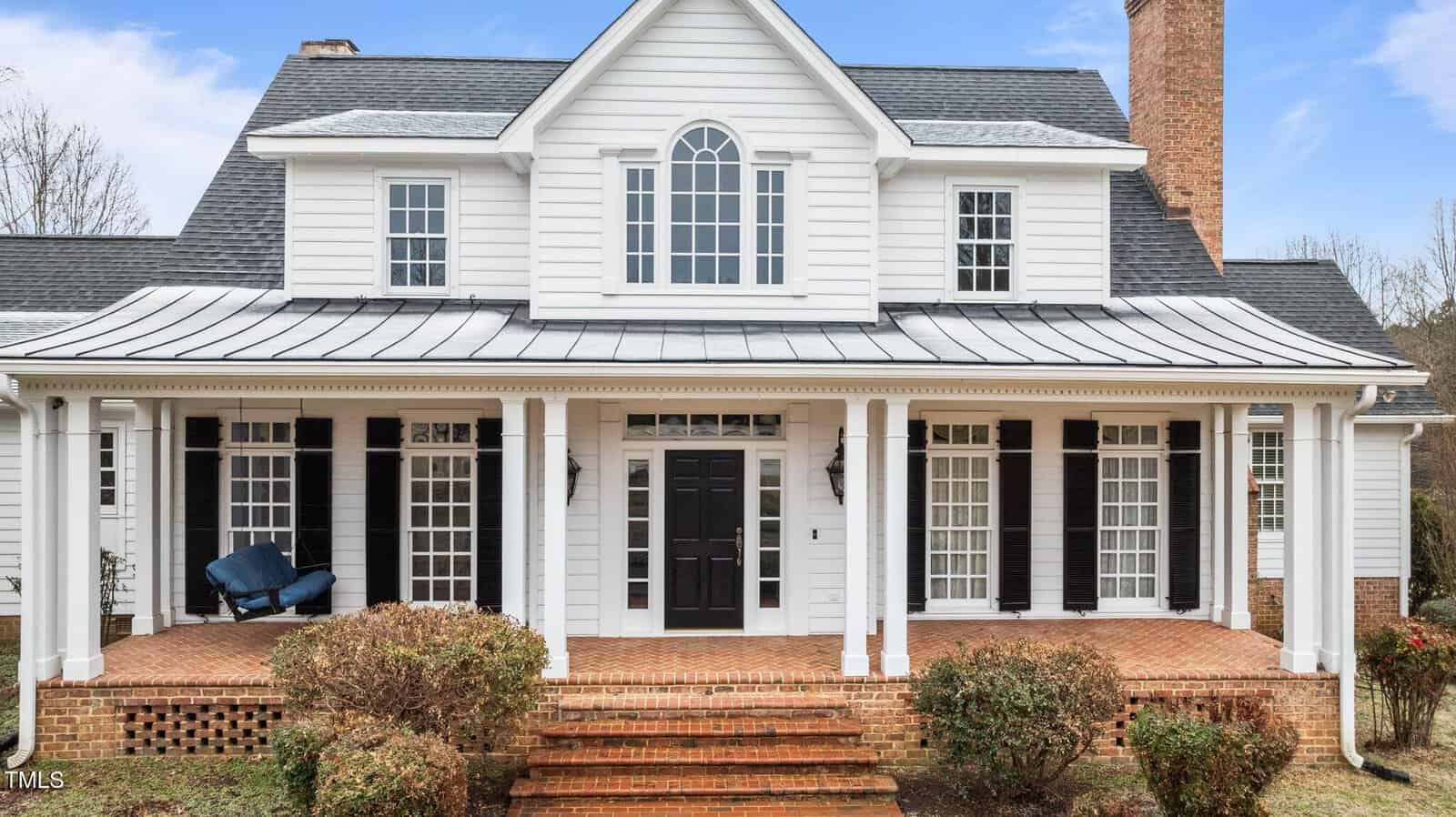 A two-story white house with black shutters, a large front porch supported by columns, brick steps, and trimmed bushes in front. The house features multiple windows and a central black front door.