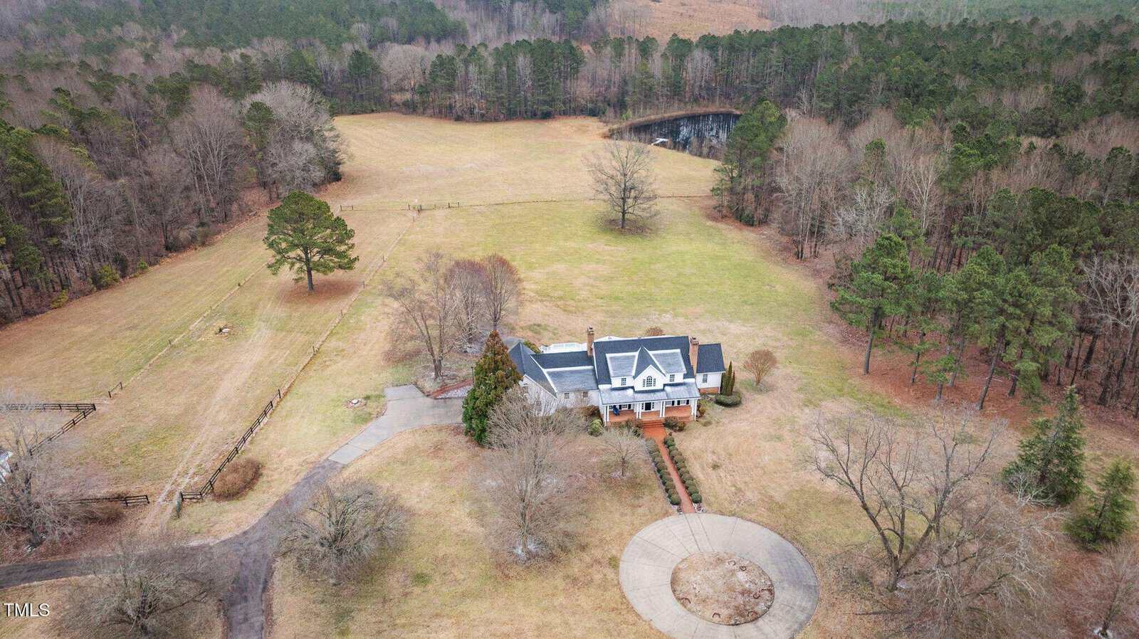 Aerial view of a large house with a covered porch, circular driveway, and spacious lawn surrounded by trees and forest, with a pond visible in the background.