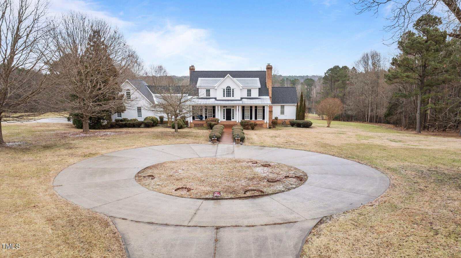 A large white two-story house with a covered front porch and twin chimneys sits behind a circular driveway, surrounded by trees and open lawn under a blue sky.