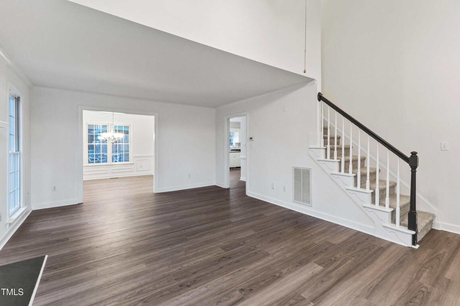 A spacious, empty room with dark wood flooring, white walls, a staircase with a dark handrail and white balusters, large windows, and an open doorway leading to another room with a chandelier.