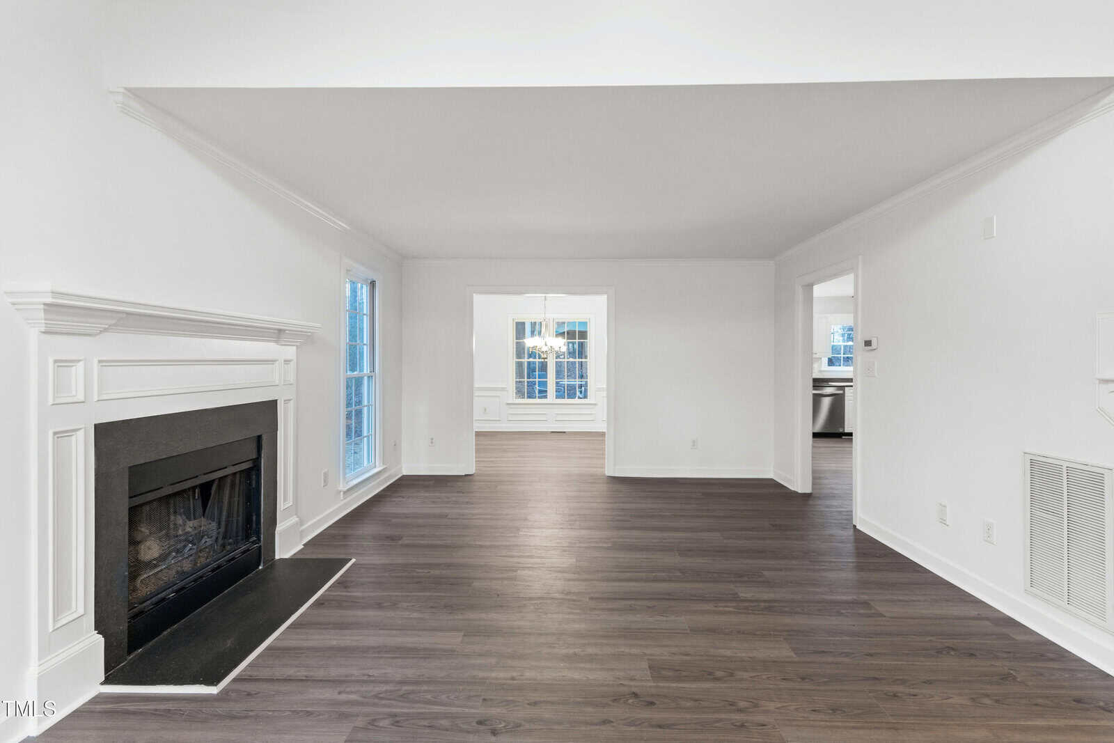 A spacious living room with white walls, dark wood flooring, a black fireplace on the left, large windows, and an open layout leading to a dining area and kitchen in the background.