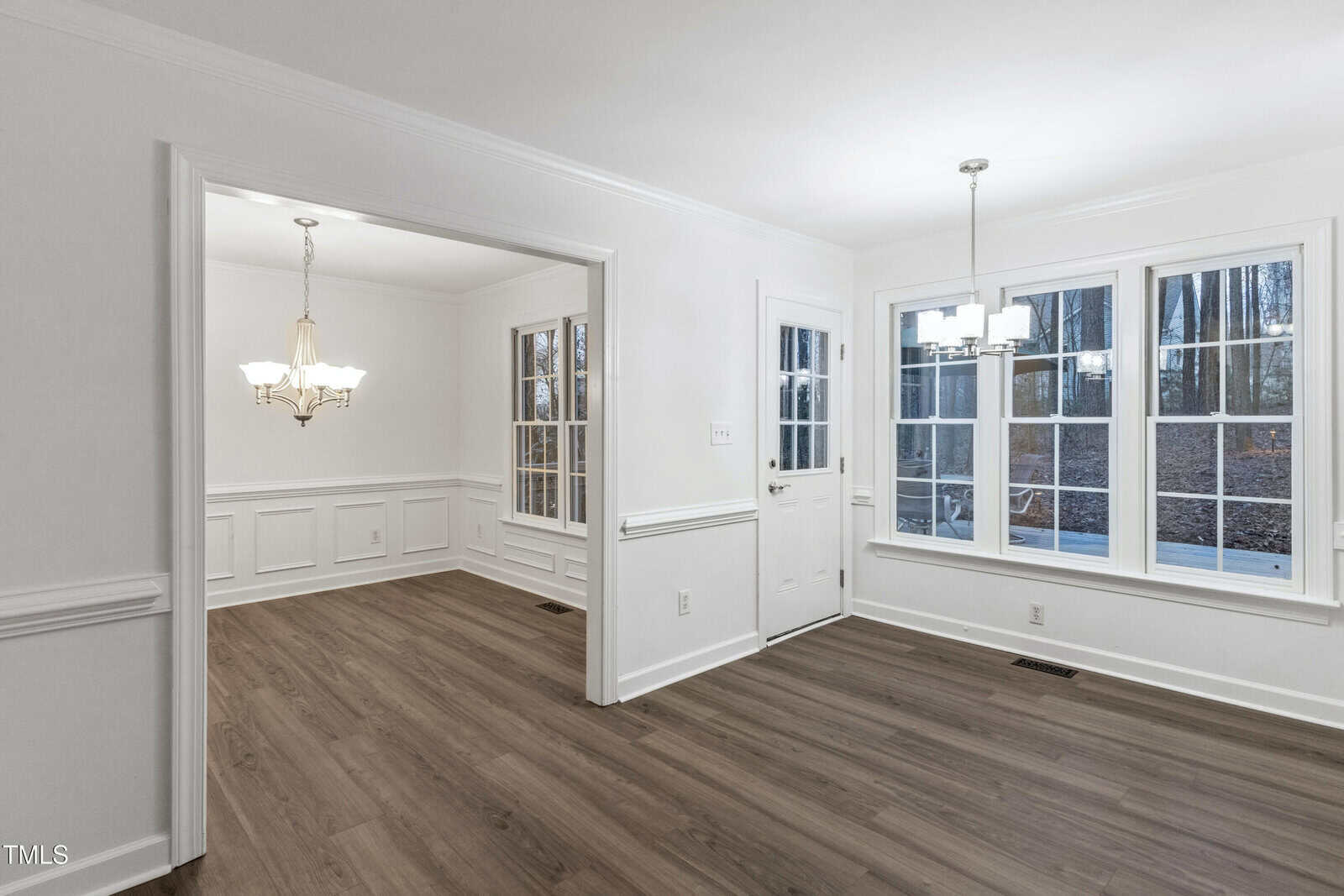 A dining and sitting area with white walls, large windows, wood flooring, crown molding, and two modern light fixtures. A door leads outside, and natural light comes through the windows.