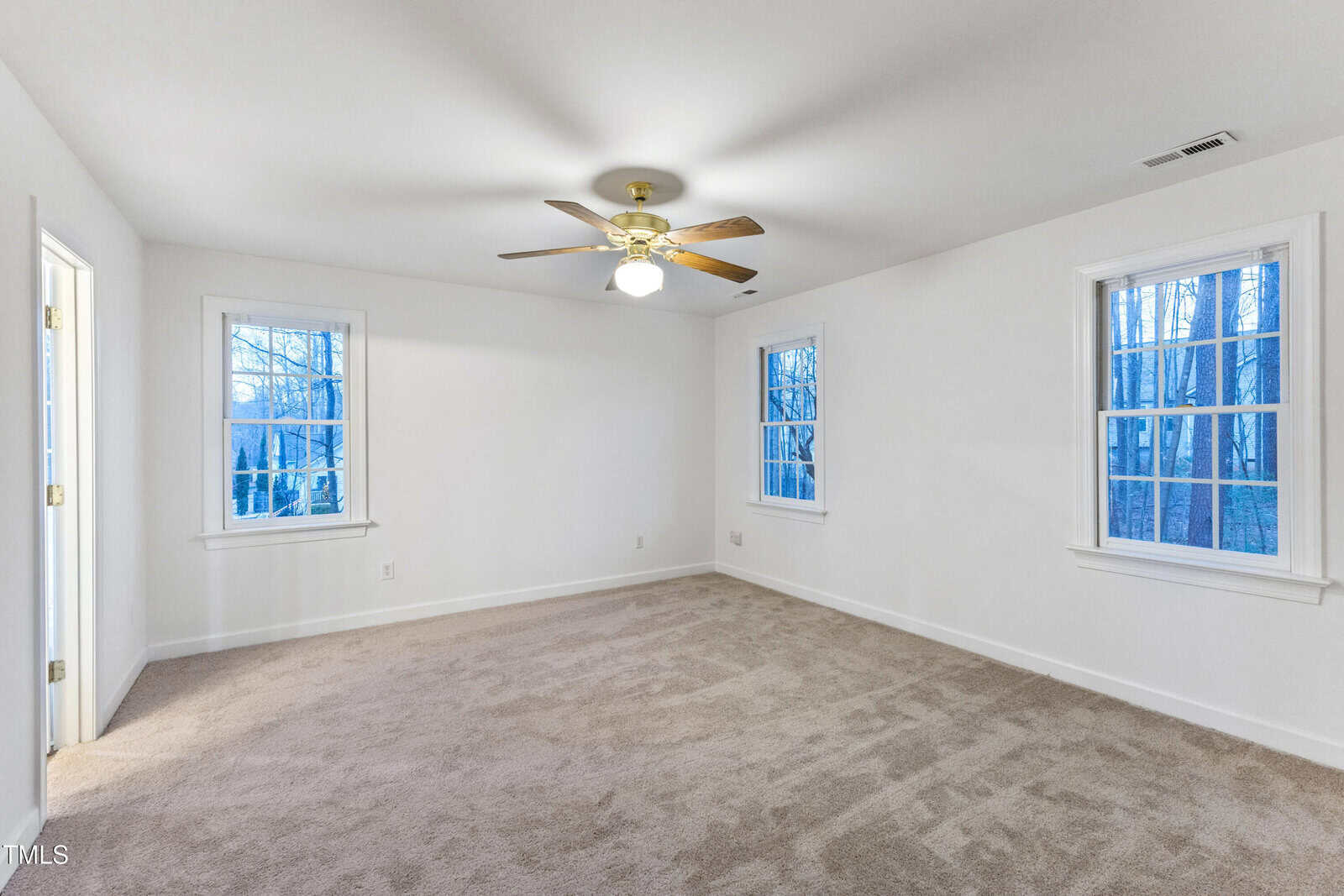 A spacious, empty bedroom with beige carpet, white walls, three windows, and a ceiling fan with a light in the center. A door to a bathroom is visible on the left side.