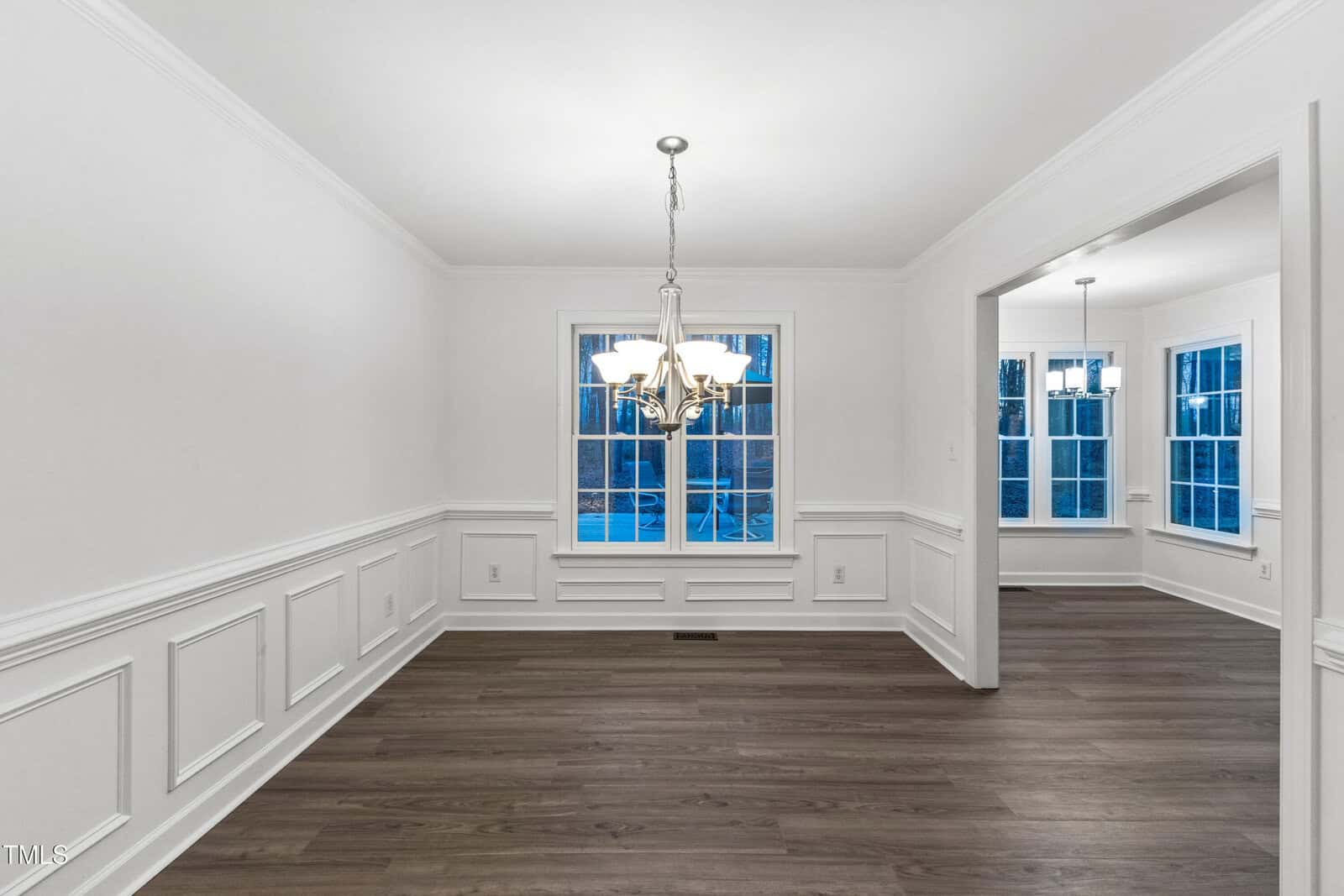 A dining room with white walls, wainscoting, dark wood flooring, a central window, and a modern chandelier. An open doorway leads to an adjoining room with similar flooring and large windows.